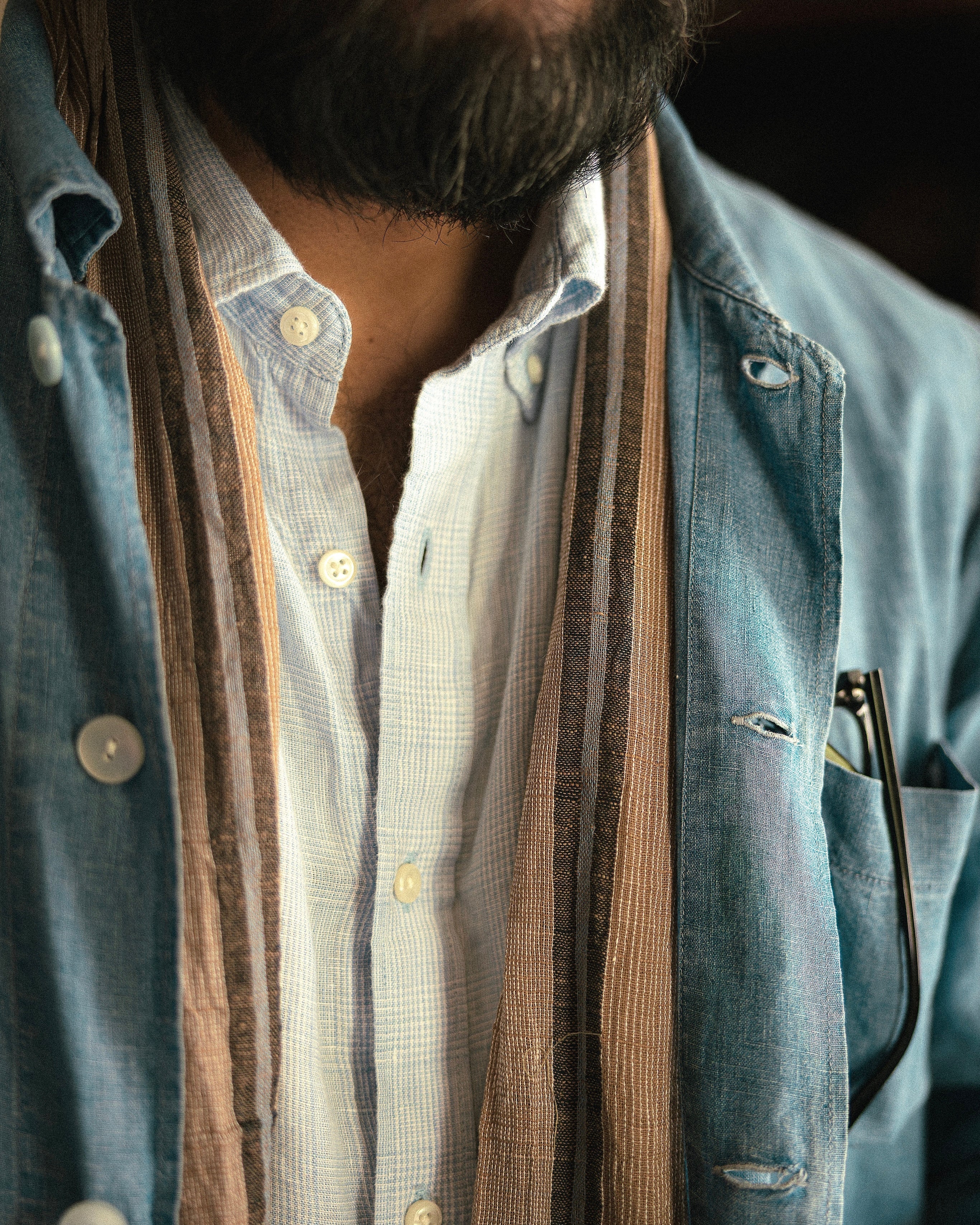 A close-up of a person wearing layered clothing, featuring a light blue denim overshirt over a white button-down shirt, with a brown and beige striped scarf draped around the neck.