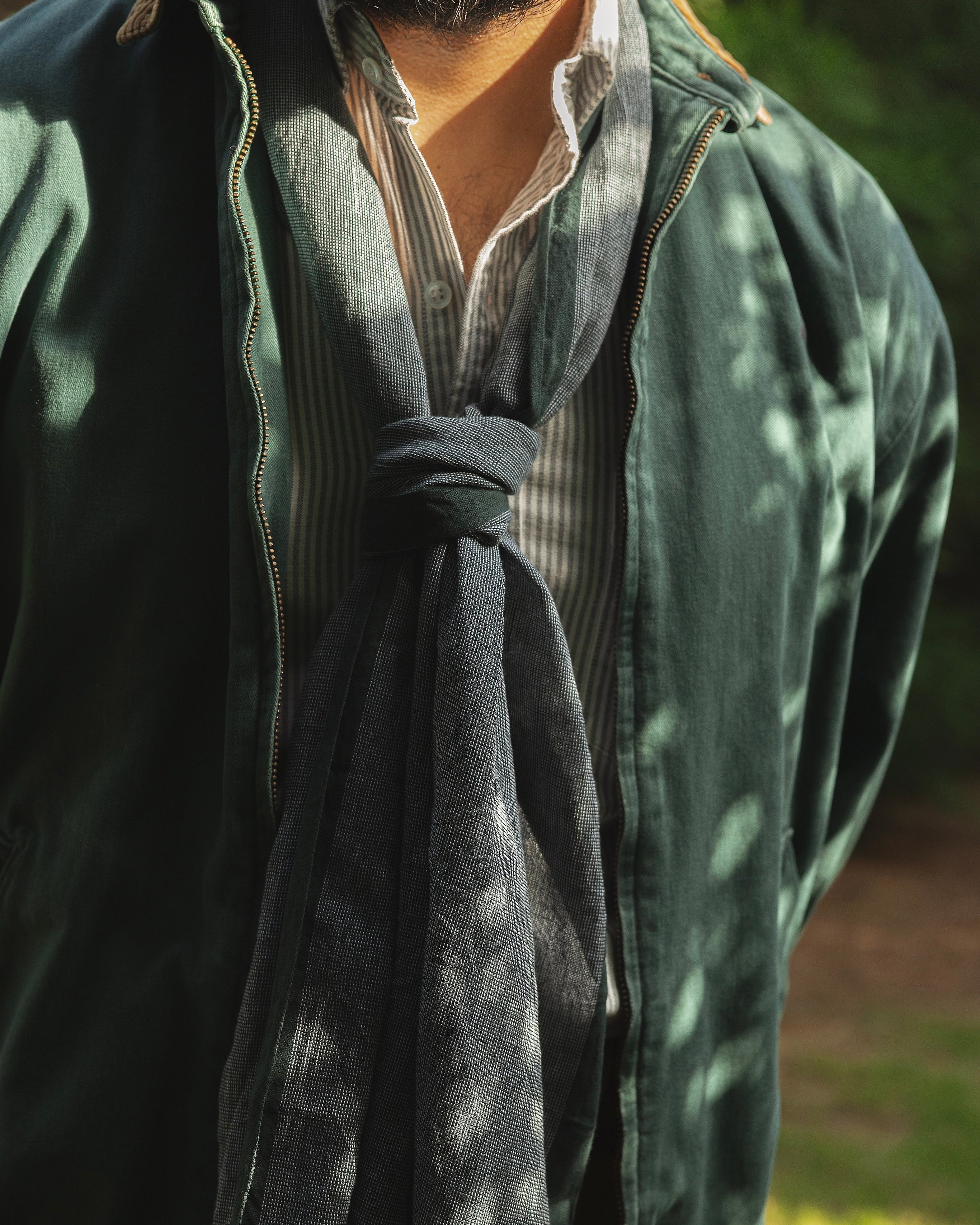 A close-up outdoor shot of a person wearing a dark green jacket over a striped shirt and a dark gray tie-neck scarf, with dappled sunlight and green foliage in the background.
