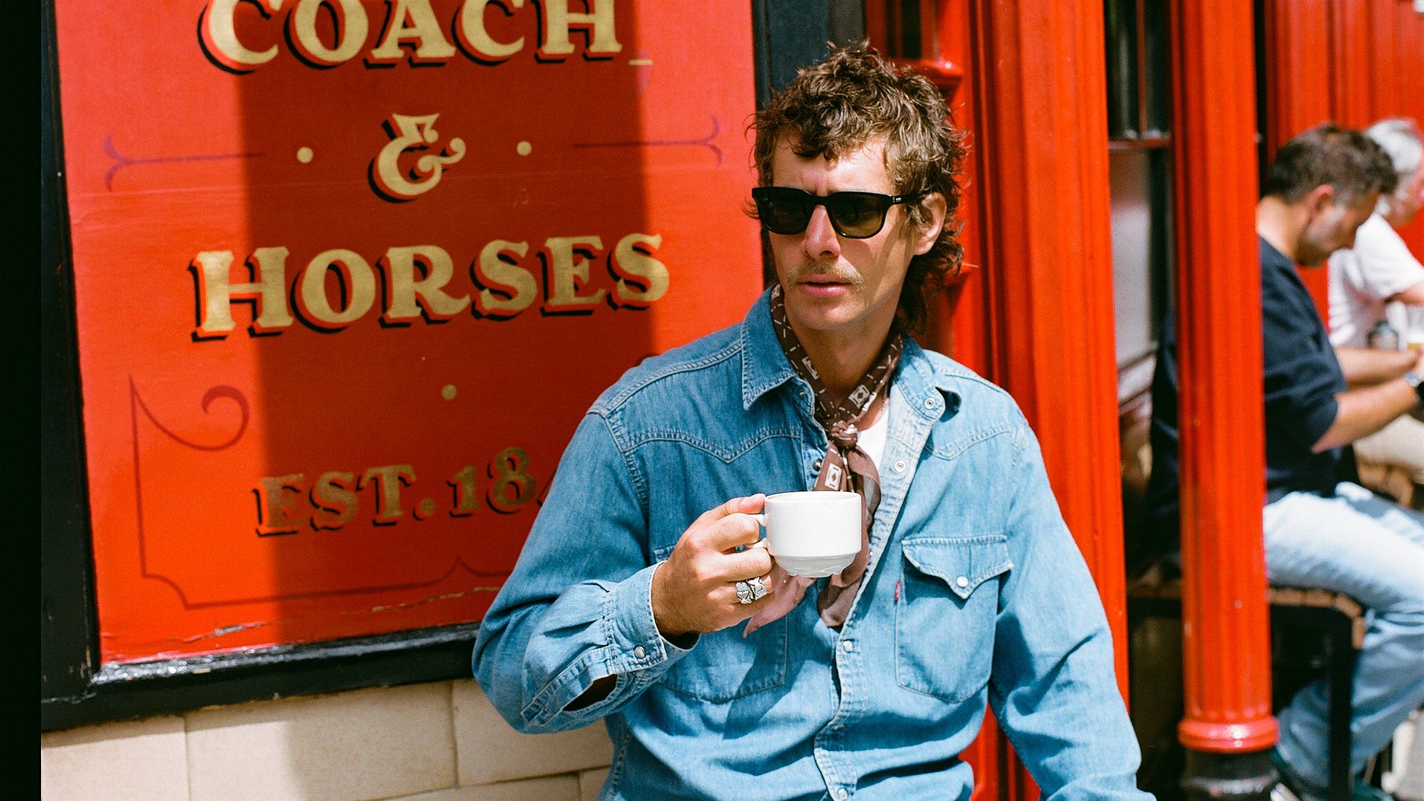 Man sitting on a bench in front of 'The Coach & Horses' pub holding a cup.
