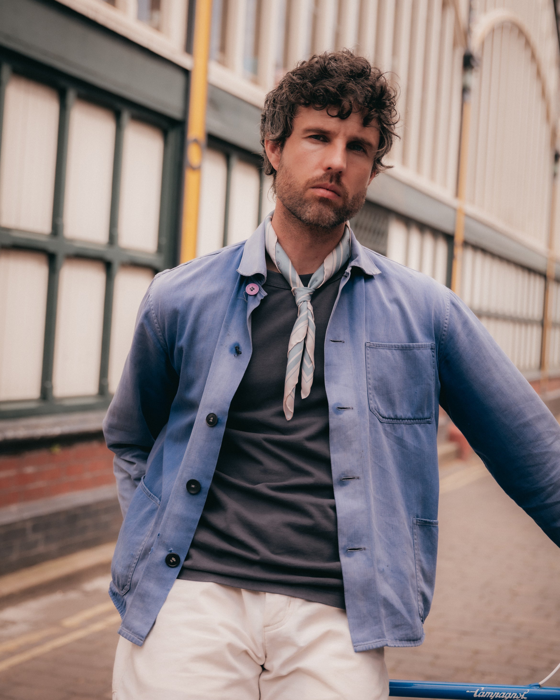 A man wearing a light blue button-up overshirt over a dark gray t-shirt, a light gray neck scarf, and off-white trousers, standing outdoors in front of a building.