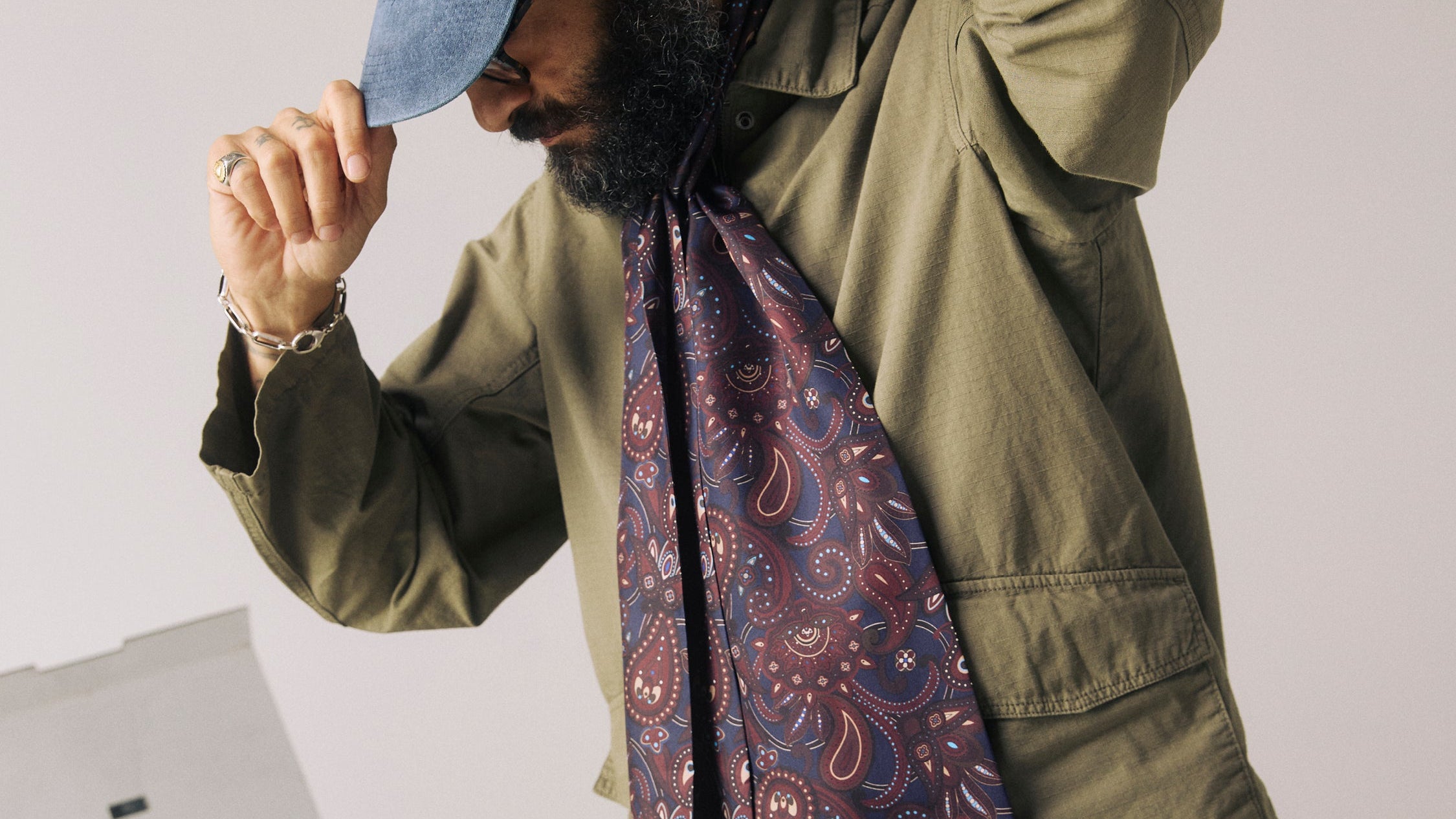 A man wearing an olive green jacket, blue cap, and a paisley patterned scarf in shades of blue, red, and white, standing in front of a light gray wall with a vinyl record player in the background.