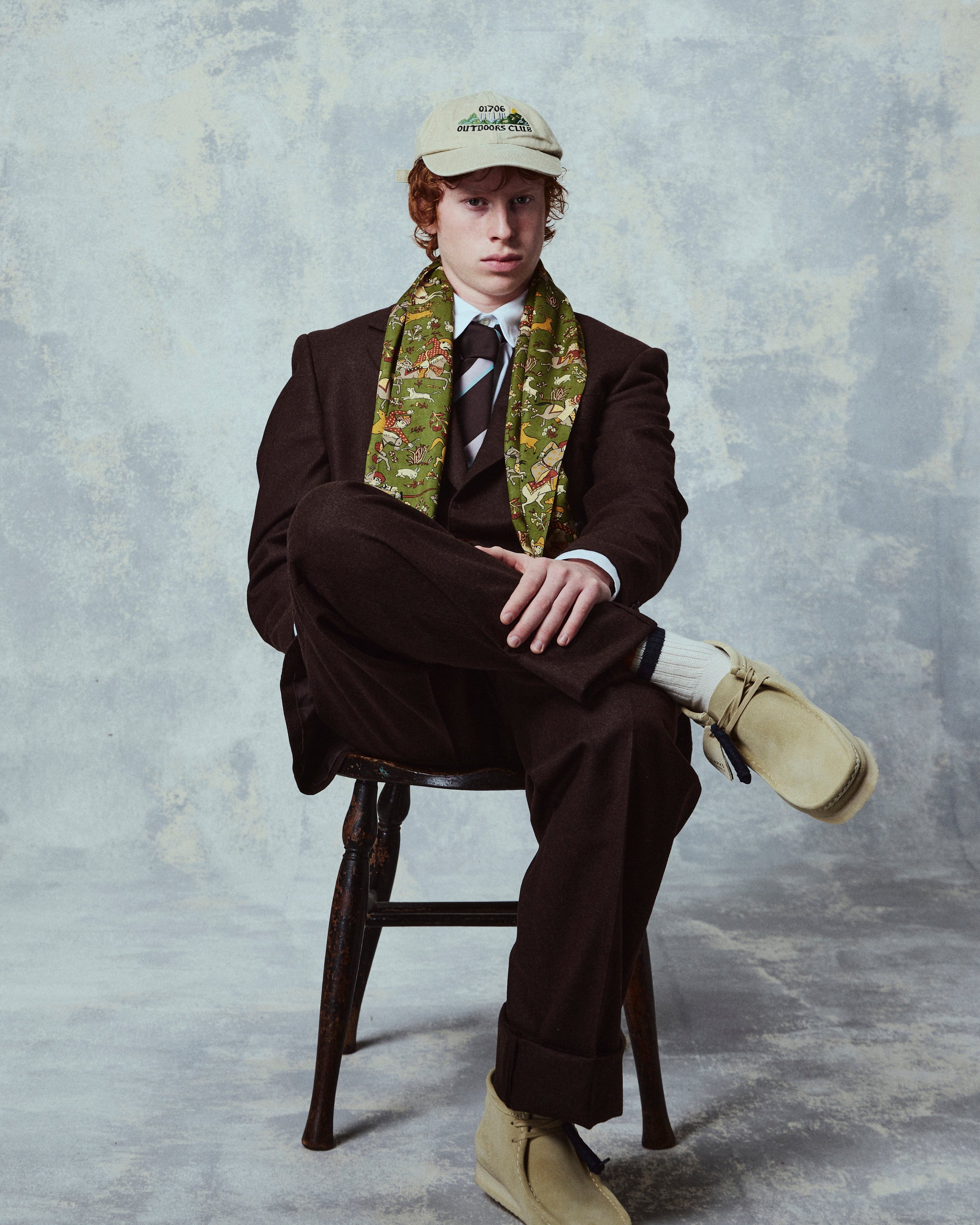 Person wearing a brown suit and beige cap sitting on a stool against a textured gray background