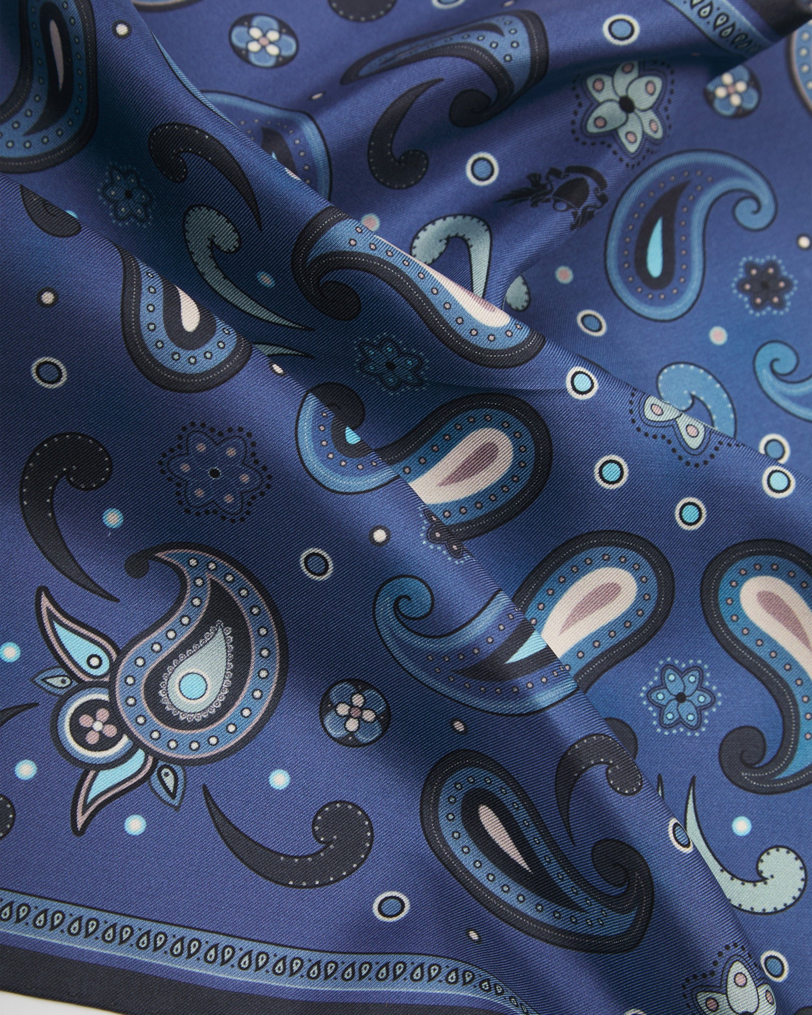 A ruffled close-up of the 'Belgravia' silk pocket square from SOHO Scarves, showing overlapping navy, sky blue and cream paisley motifs on a rich cobalt-blue ground, accented with delicate floral rosettes and fine circle dots.