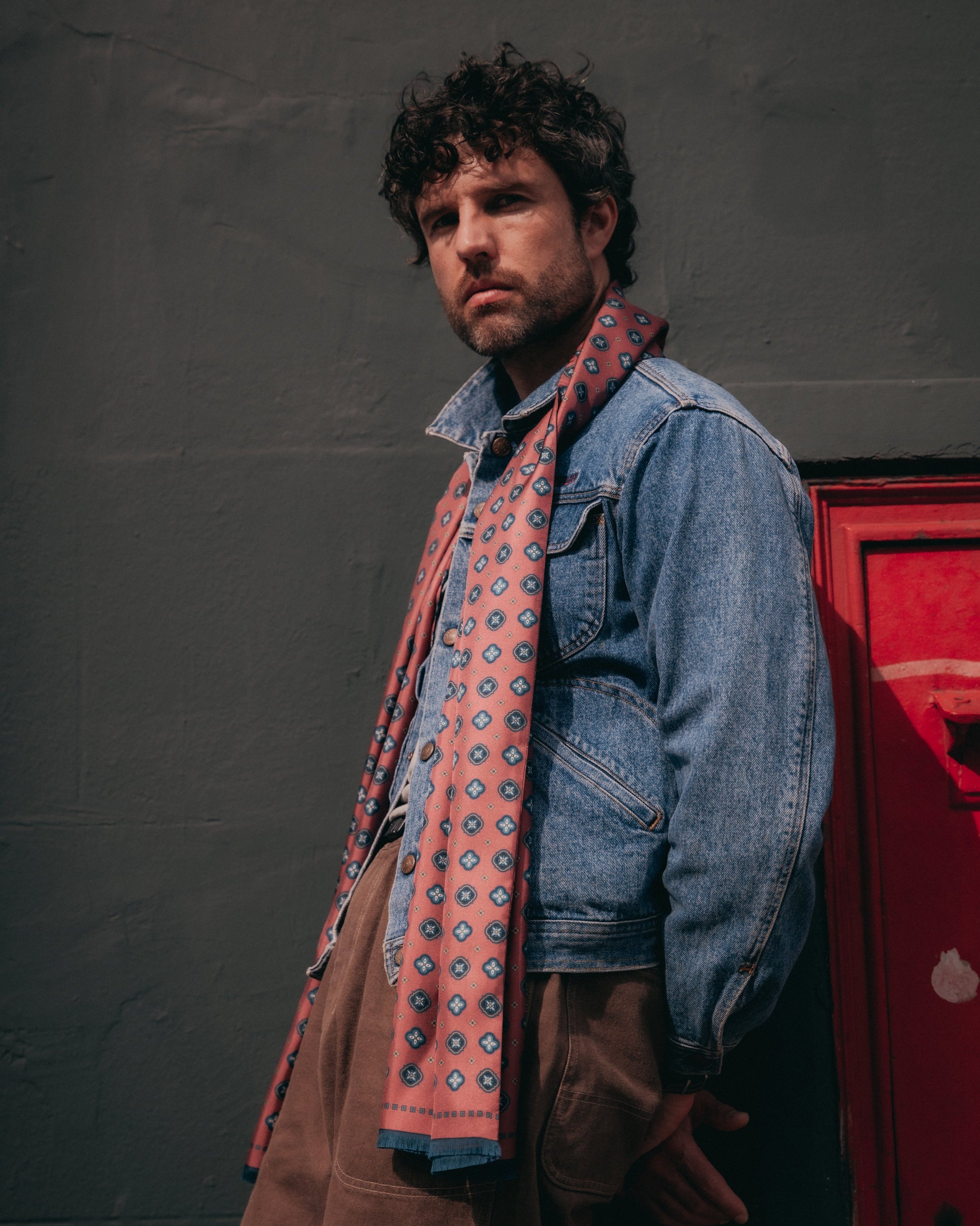 Man standing with arms apart, leaning on a wall wearing the berry red 'Chelsea' silk scarf loosely around the neck and paired with brown denim pants and a denim jacket.