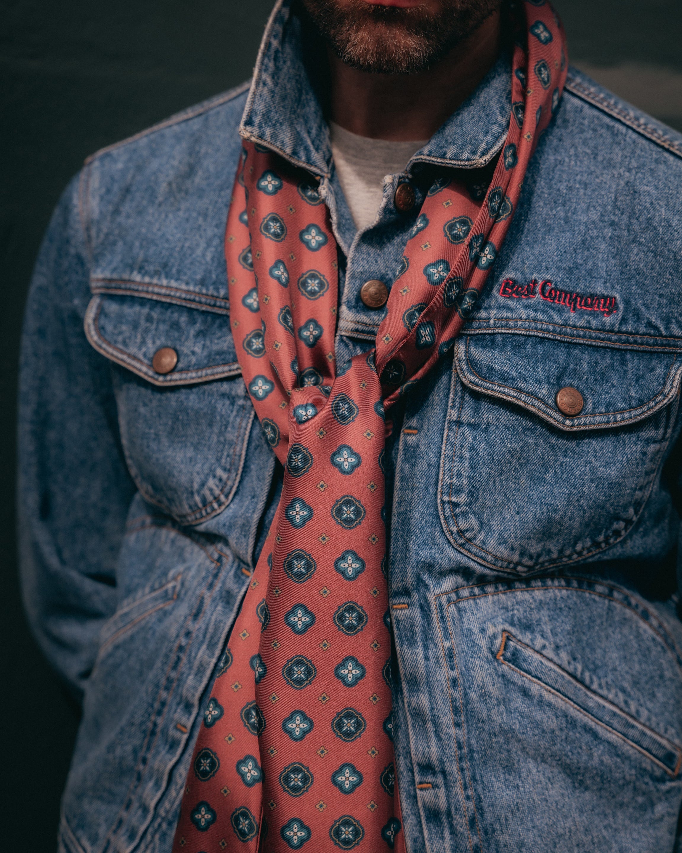 Close-up view of man wearing the berry red 'Chelsea' silk scarf from SOHO Scarves with a clear view of the patterns comprising of pale teal cross motifs and circle medallions.