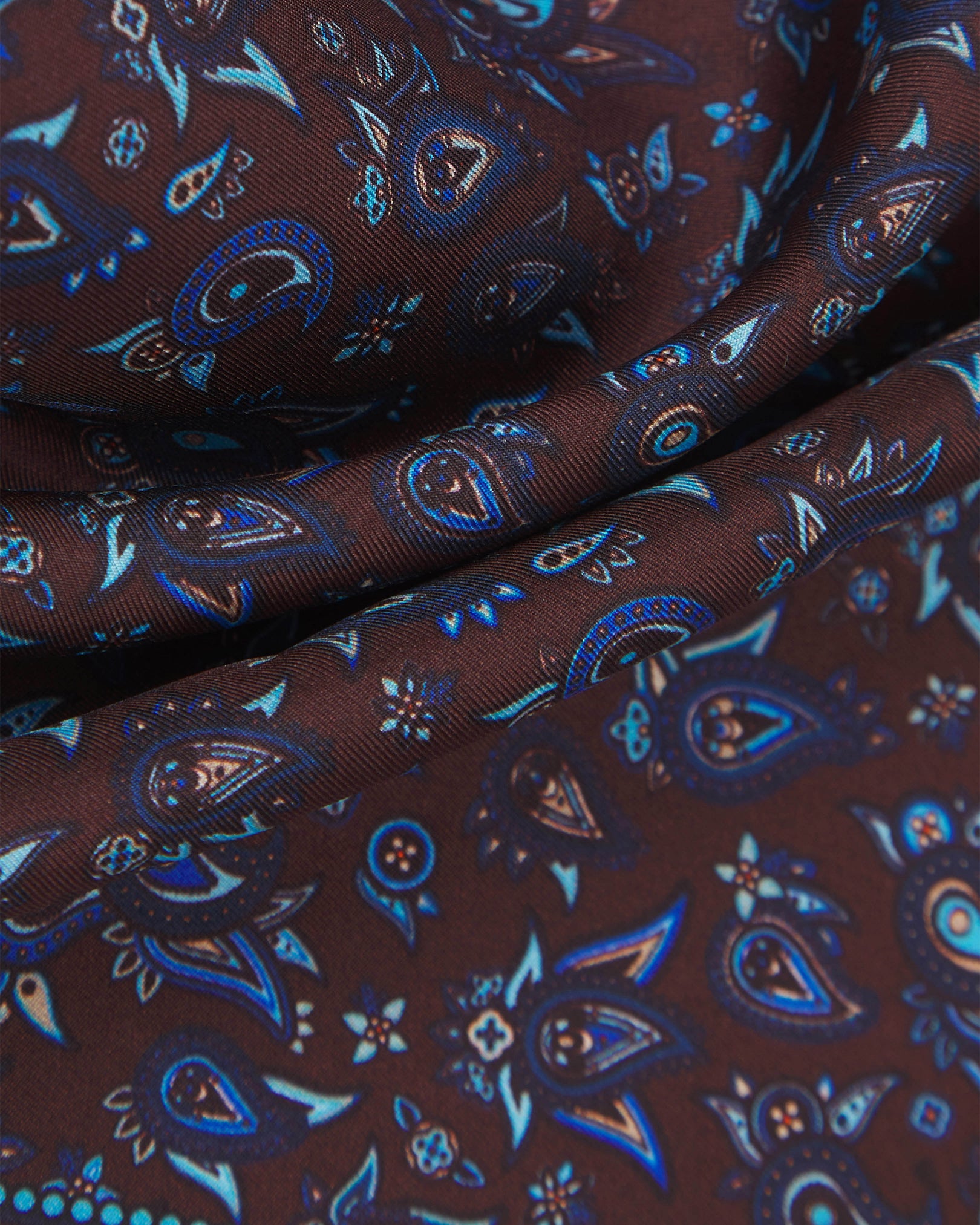 A ruffled close-up of the 'Buttermere' silk pocket square from SOHO Scarves, showing overlapping elements of the paisley decoration against a brown-plum background.