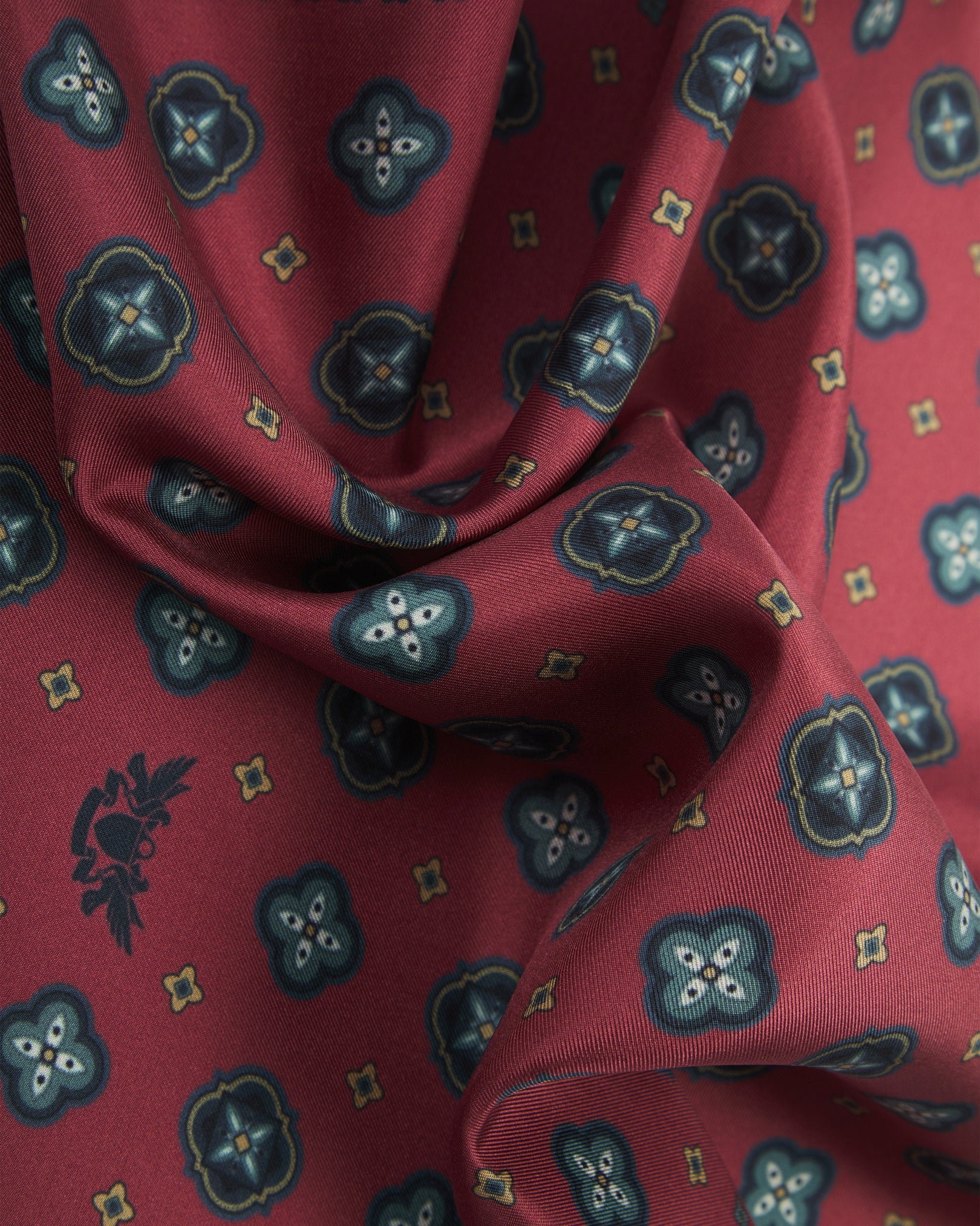 A ruffled close-up of the 'Chelsea' silk pocket square from SOHO Scarves, showing overlapping teal quatrefoil medallions with white star motifs and fine gold diamond accents on a deep crimson-berry ground.