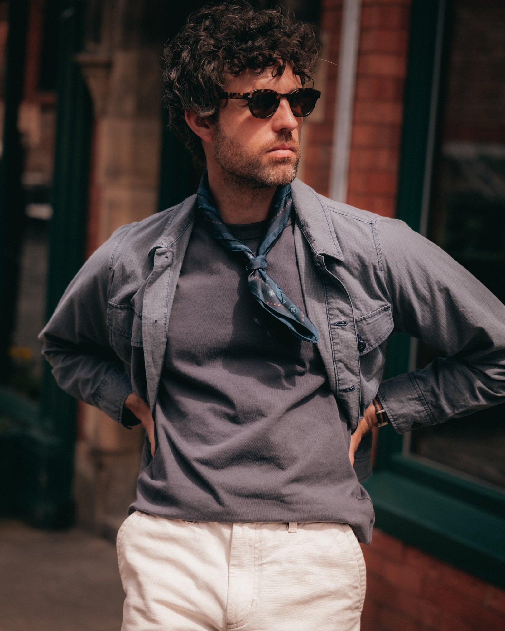 A man wearing a grey button-up shirt over a dark gray t-shirt, cream pants, and sunglasses, standing outdoors with his hands on his hips wearing the silk 'Chelsea' indigo neckerchief.