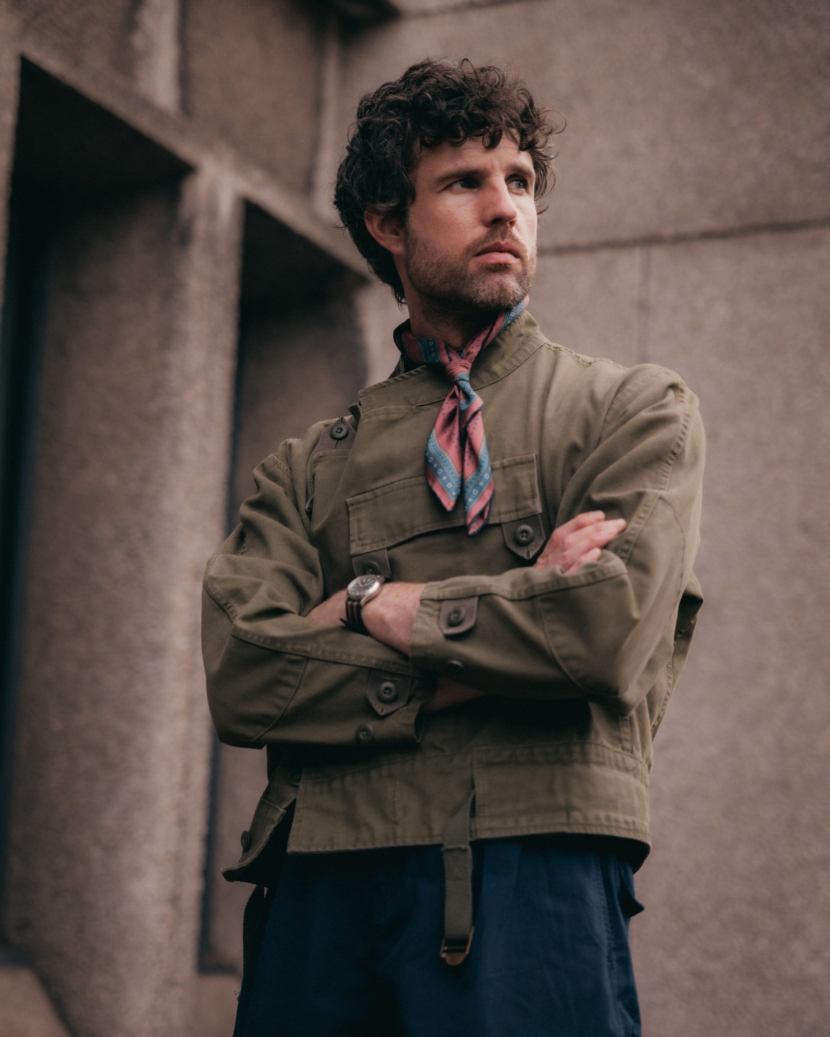 A man wearing a brownish-green utility jacket, the 'Chiswick in Berry' silk neckerchief and dark baggy shorts standing with arms crossed.