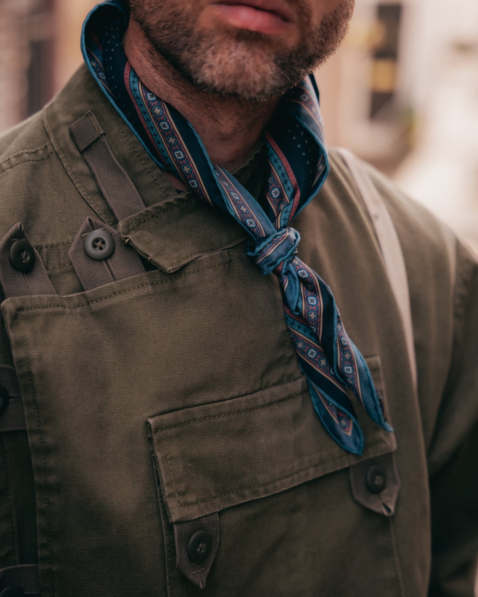 A close-up lifestyle shot of a man wearing a dark olive green utility jacket and the indigo silk neckerchief from SOHO Scarves.