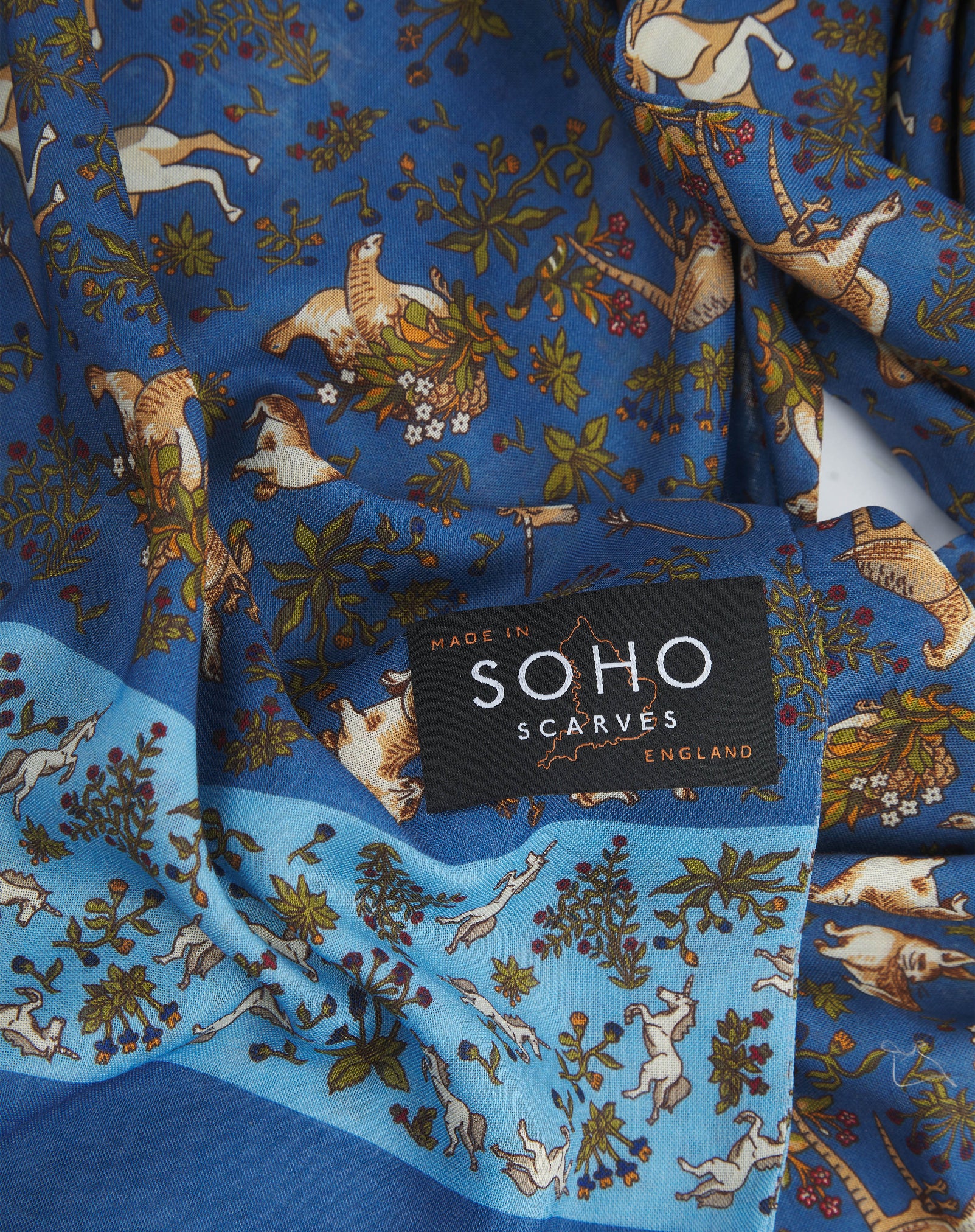 A ruffled, close-up view of the UK-made blue-blue silk and wool scarf depicting unicorns, hares and pheasants amid botanical forms. Clearly showing the premium silk and wool weave against a blue background.