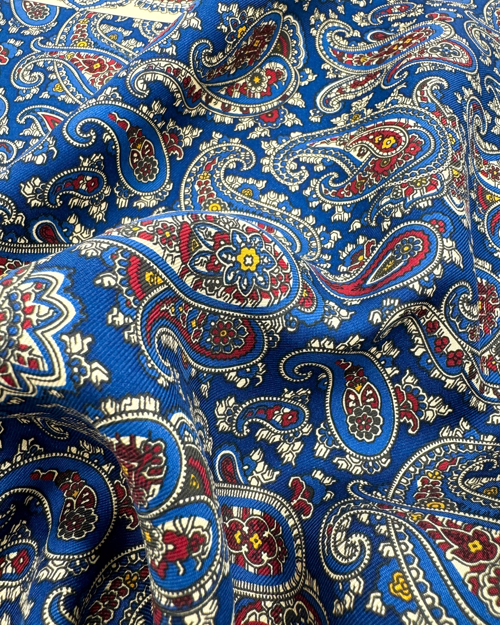 A ruffled close-up of the 'Abbotsbury' pocket square, presenting a closer view of the blue, red and cream paisley patterns against the attractive lustre of the madder silk material. 