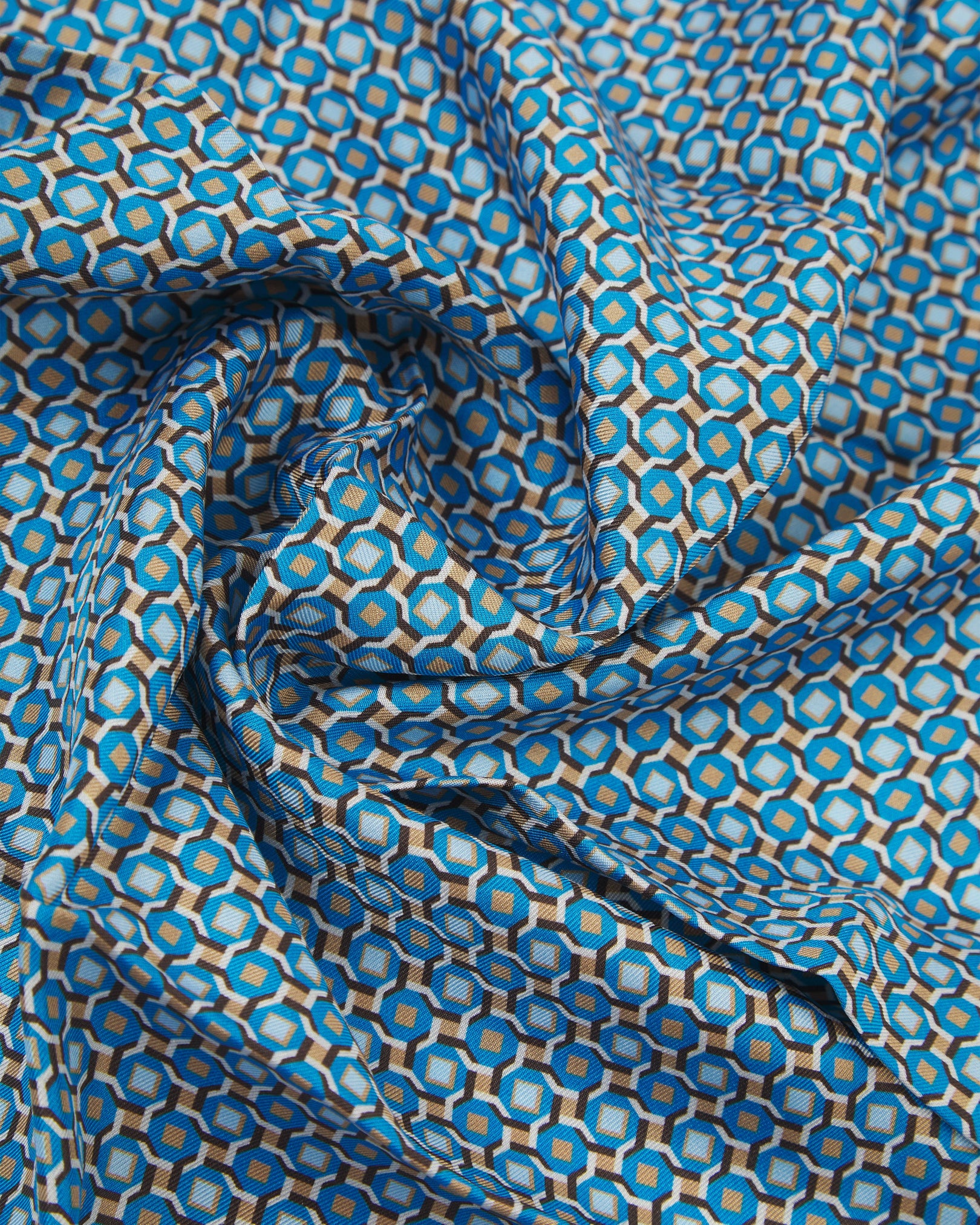 A ruffled close-up of the 'Berwick' silk neckerchief from SOHO Scarves, showing interlocking hexagonal shapes in turquoise, stone-grey, and gold with a fine, textured weave.