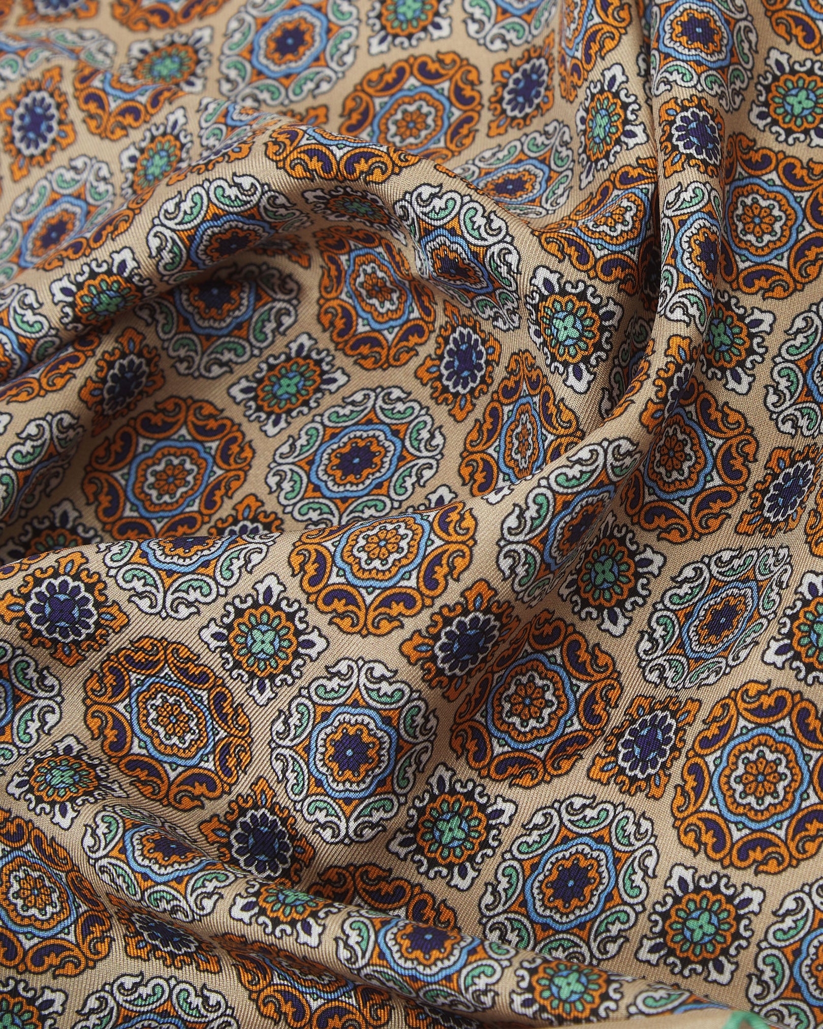 A ruffled close-up of the 'Fitzrovia' silk neckerchief from SOHO Scarves, highlighting the intricate medallion pattern in blue, green, and ochre tones set against the soft hazelnut silk.
