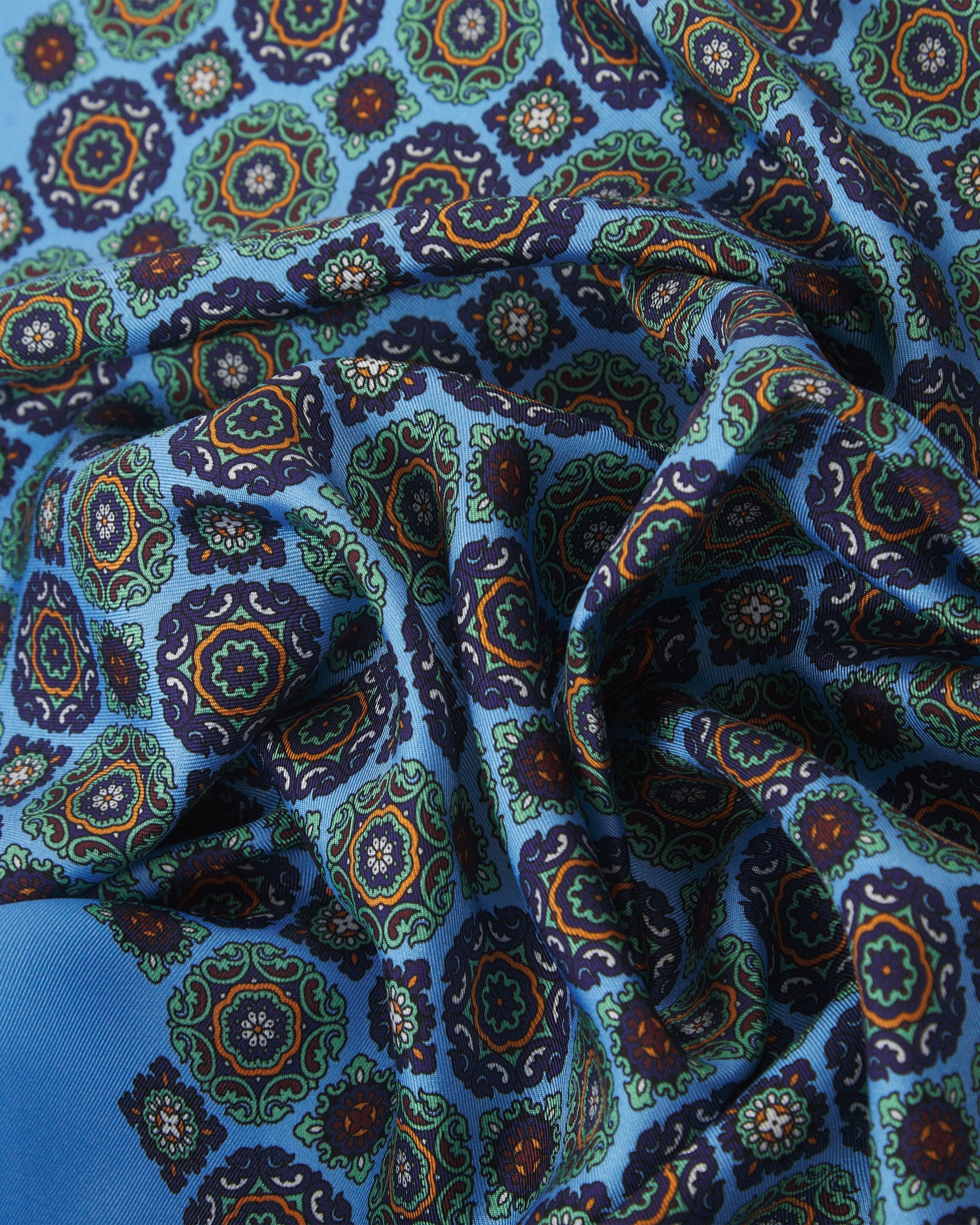 A ruffled close-up of the 'Fitzrovia' silk neckerchief from SOHO Scarves, showcasing the detailed floral medallions in deep navy, green, and red tones on a textured blue background.