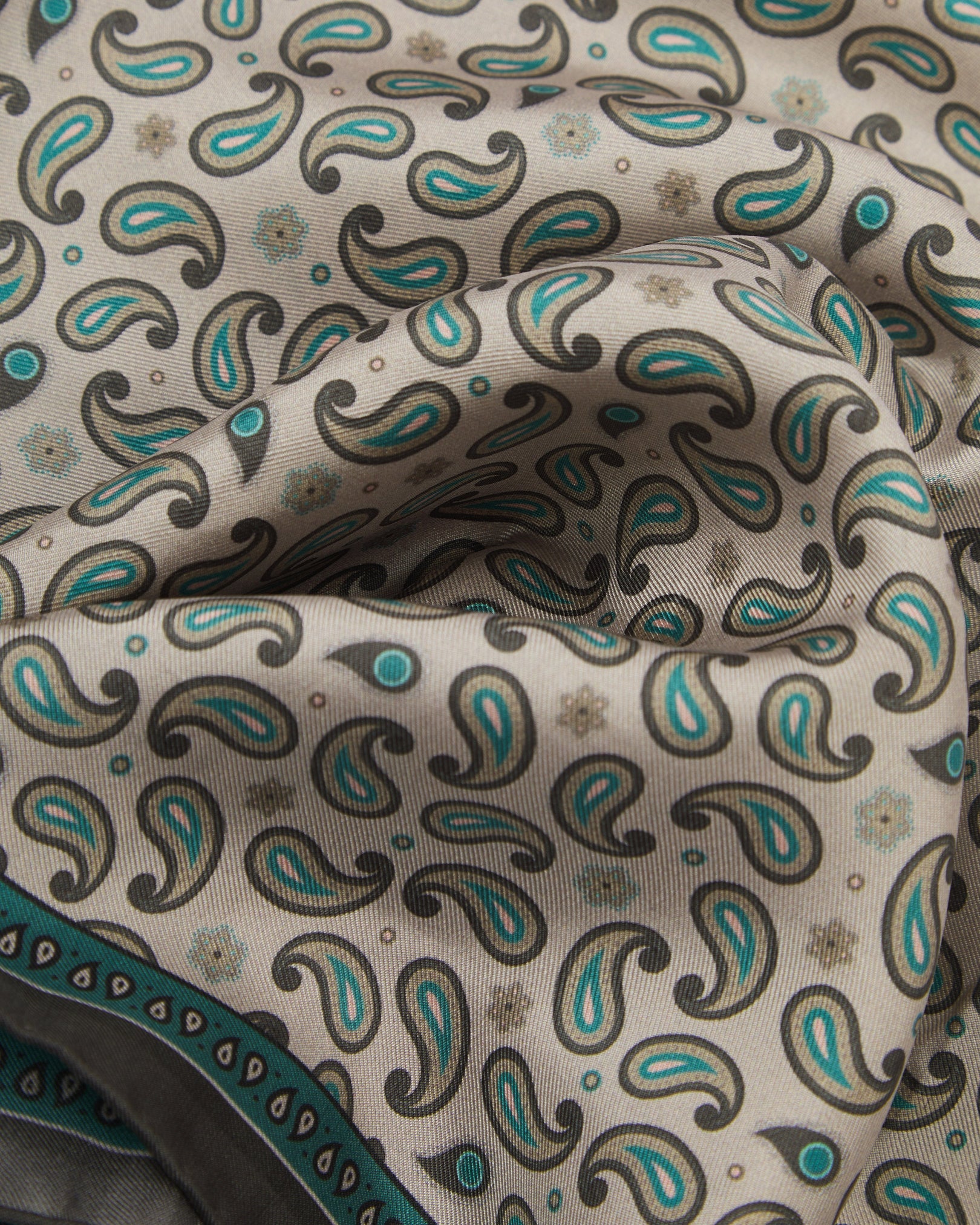 A ruffled close-up of the 'Highbury' silk neckerchief from SOHO Scarves, featuring a delicate repeating paisley pattern on a warm khaki ground with a finely woven silk texture, accented by teal and olive detailing.