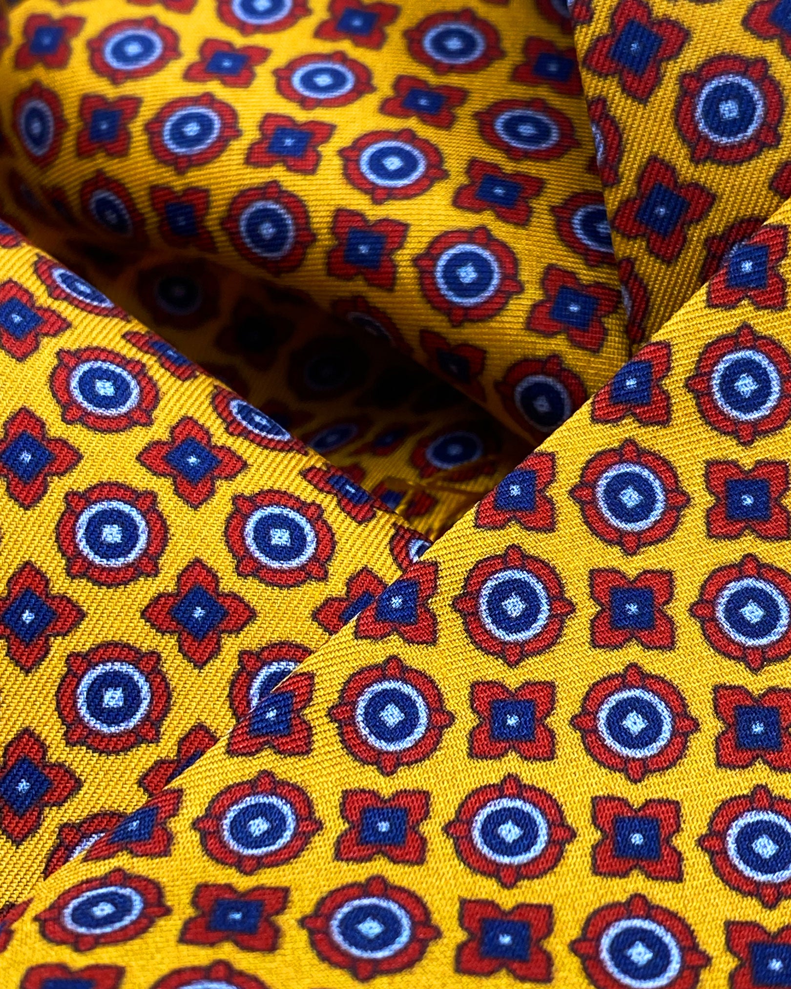 Ruffled close-up view of the Toshima silk aviator scarf, presenting a closer view of the small blue and red stylised floral patterns on a golden-orange ground and lustre of the material.