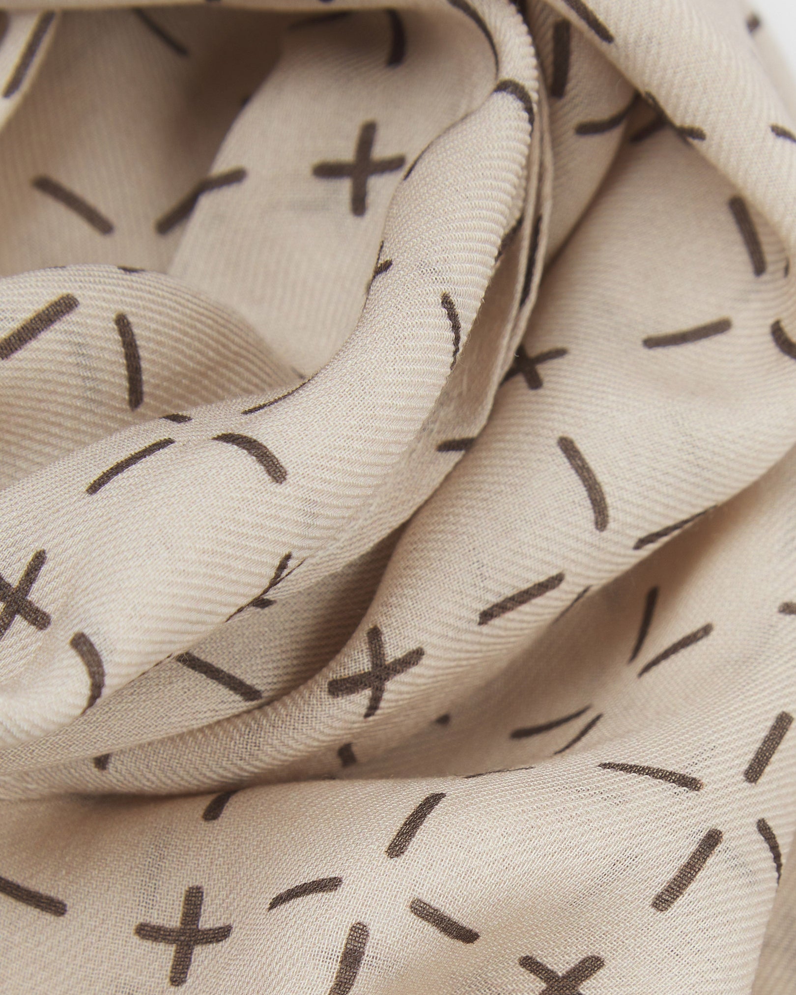 A ruffled close-up of the 'Crosses' cream bohemian wide scarf, showing the soft modal and wool blend fabric with a repeating geometric grid of dark grey dashes and crosses on a pale cream background.