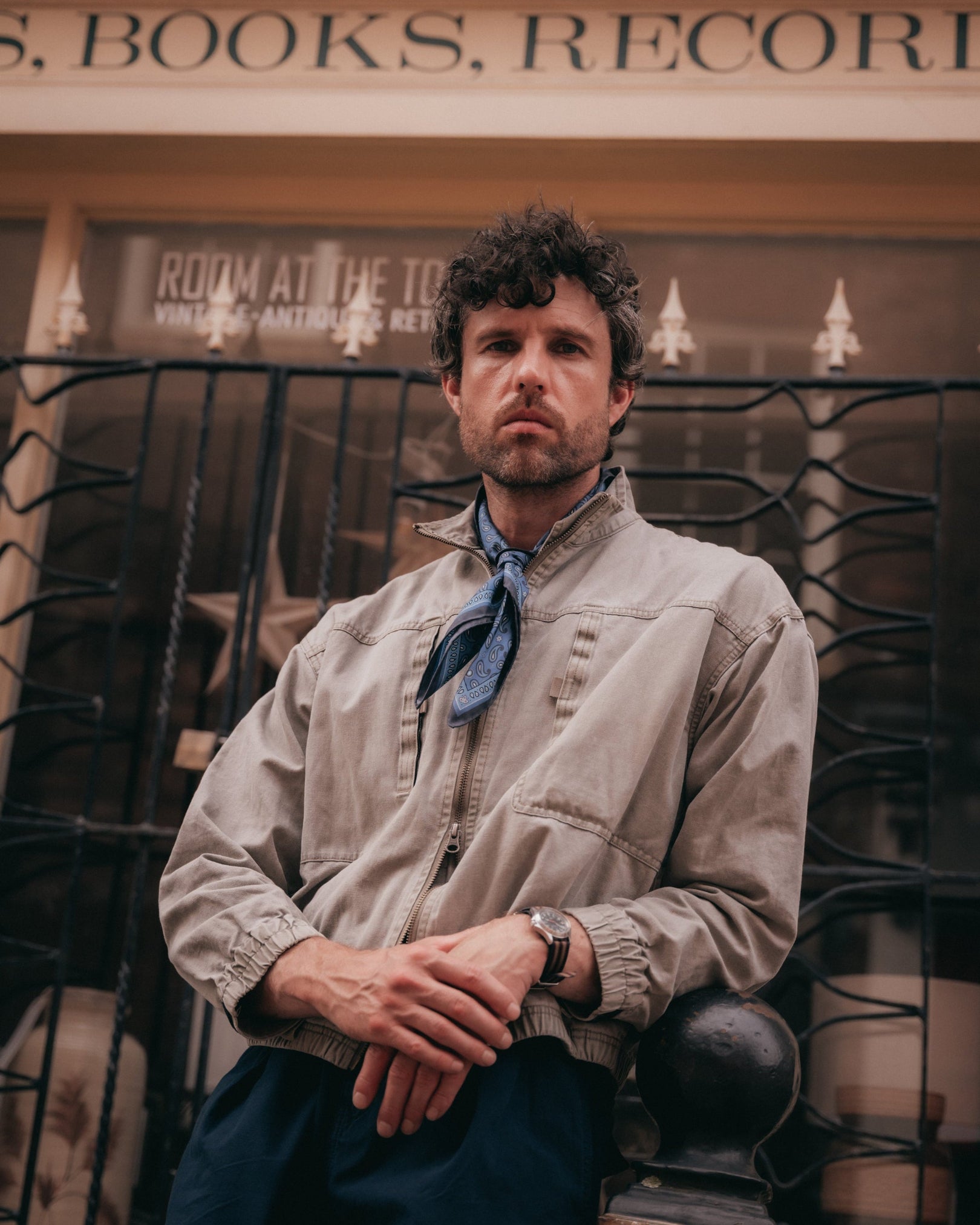 A man leaning against some railings outside a record shop wearing a light beige utility jacket with the 'Highbury in cornflower Blue' silk neckerchief.