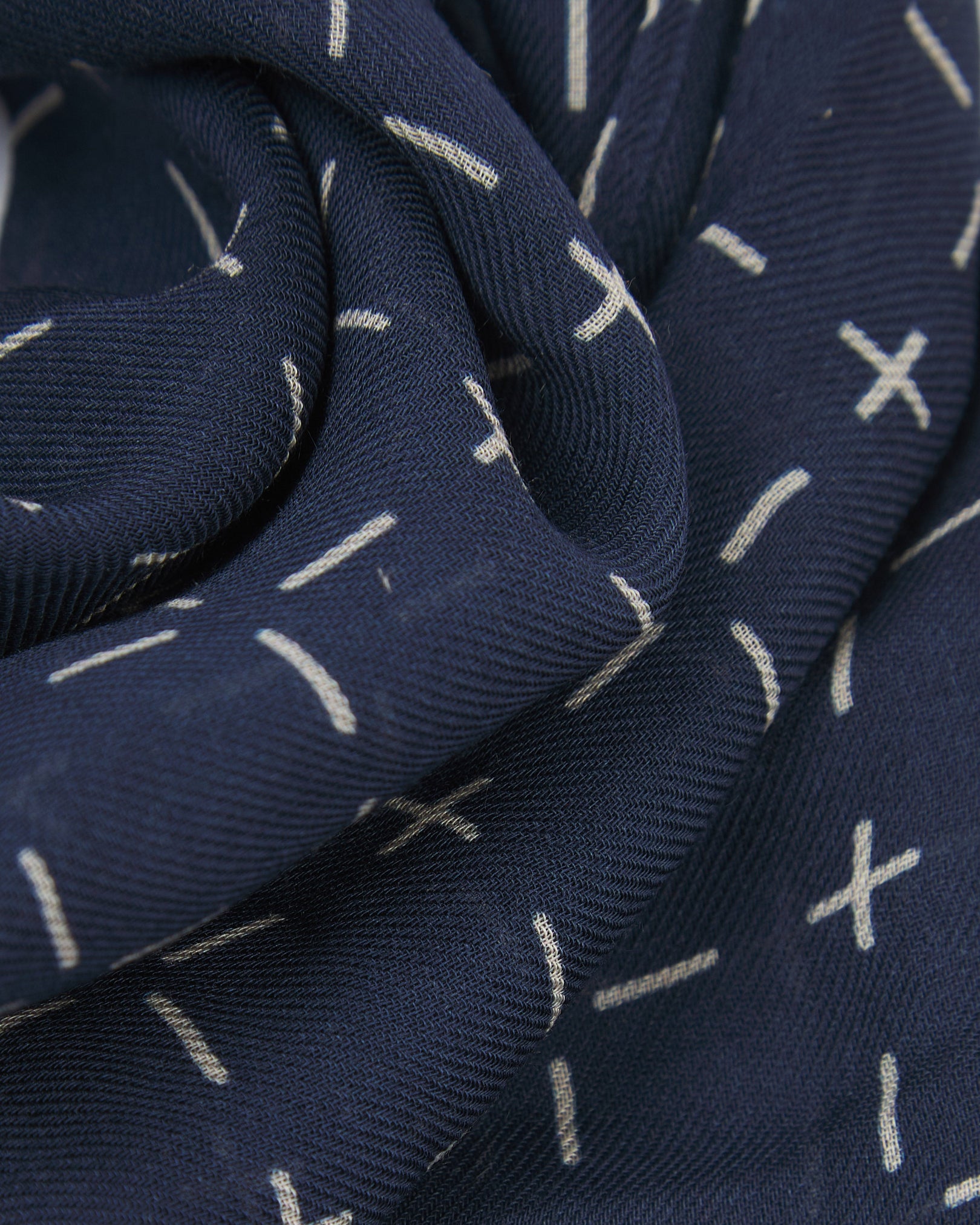 A ruffled close-up of the 'Crosses' indigo bohemian wide scarf, showing the soft modal and wool blend fabric with a repeating geometric grid of white dashes and crosses on an indigo blue background.