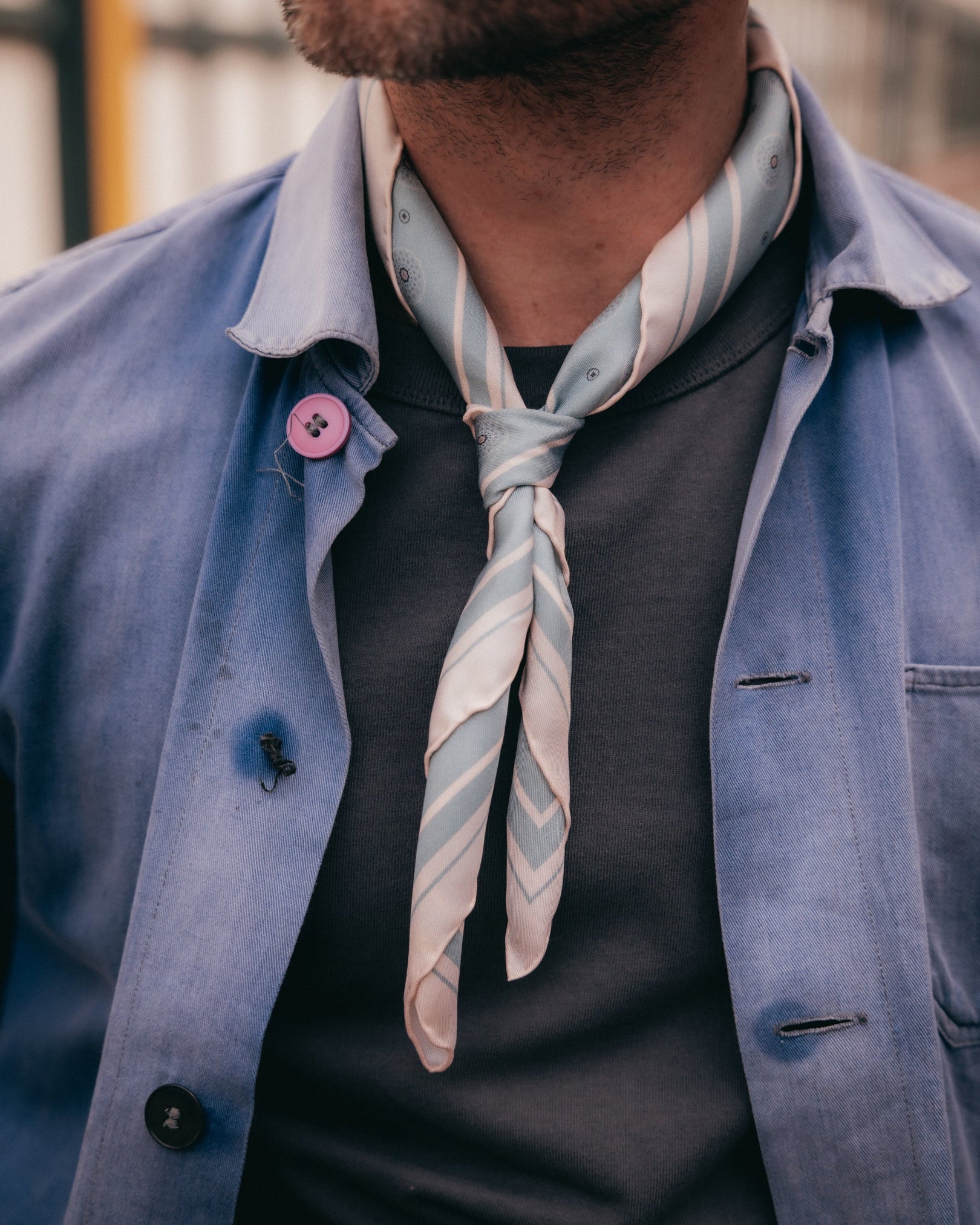 A close-up of a man wearing a light blue striped neckerchief tied around his neck, layered over a dark gray t-shirt and a light blue button-up shirt.