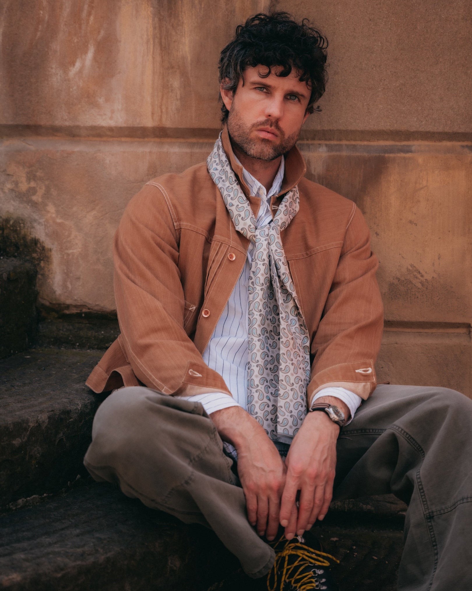 Man sitting on steps wearing the pale khaki 'Highbury' silk scarf loosely around the neck.