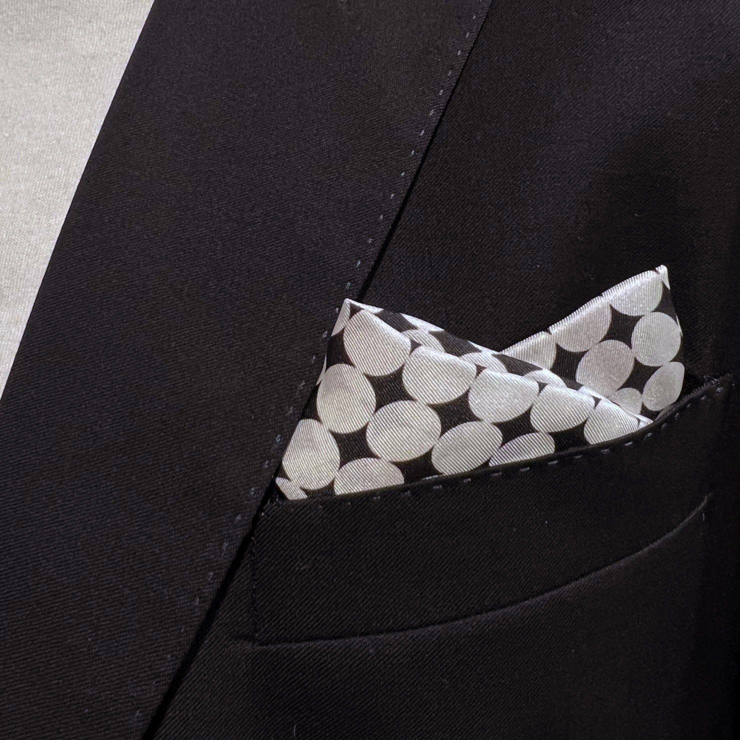 Close-up of 'Keaton' silk pocket square with white disc patterns in breast pocket of dark suit jacket.