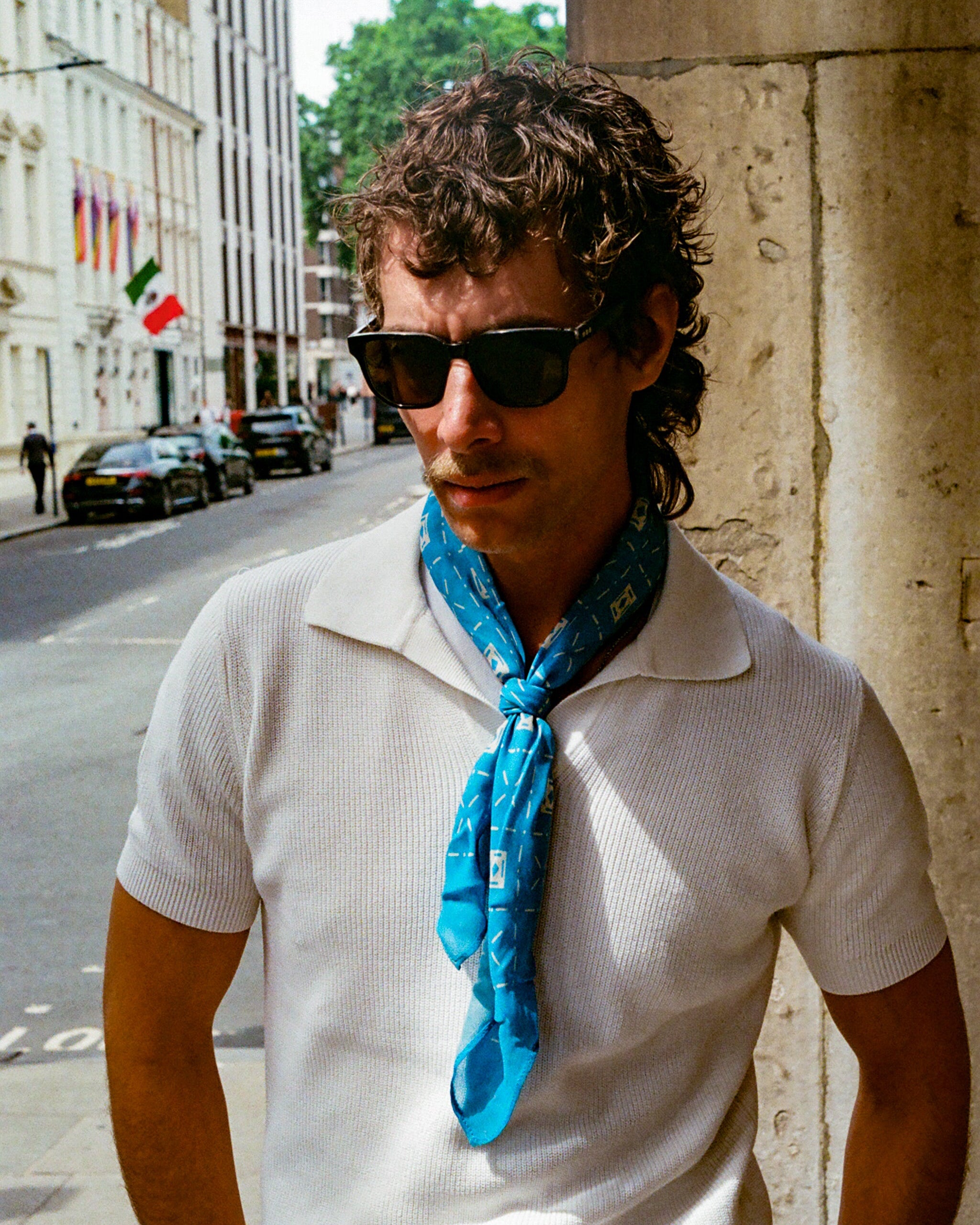 A man wearing the 'Diamonds in Pacific' modal and wool bandana from SOHO Scarves, loosely tied around the neck with a white shirt, showcasing the ordered diamond mosaic pattern in contrasting Pacific blue tones.