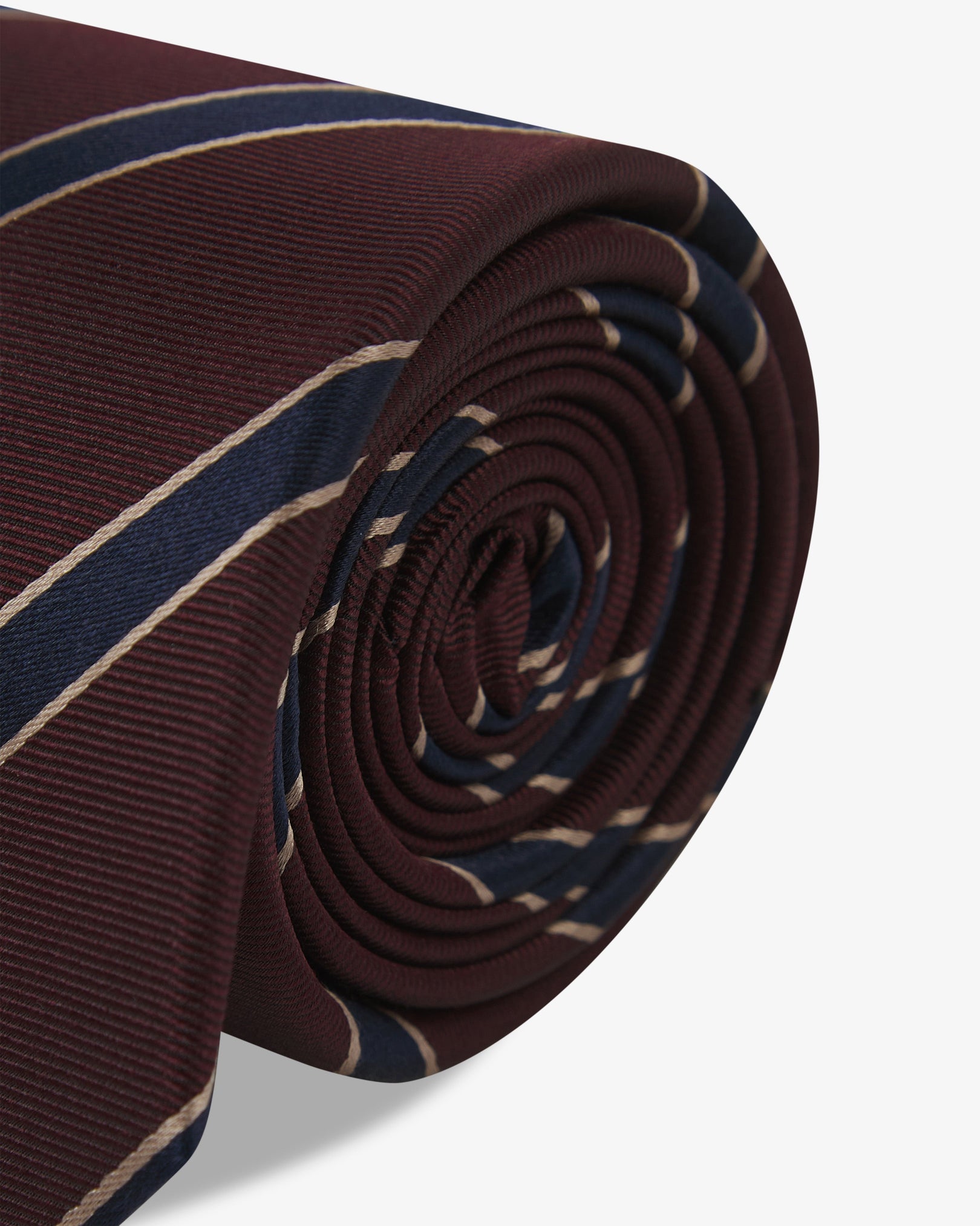 The 'Sanders Mahogany Haze' silk neck tie neatly coiled with a clear view of the silk weave and gold and navy stripes against a maroon background.