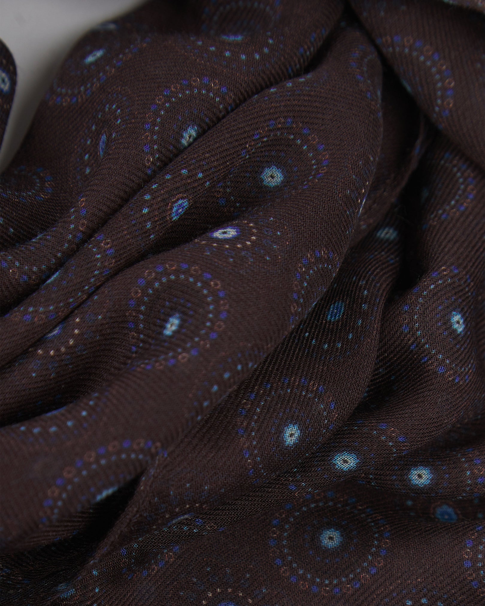 A ruffled close-up of the Seattle 'Mahogany Daze' bohemian wide scarf, showing the soft modal and wool blend fabric with intricate circular motifs in blue and brown on a dark plum background with delicate cream accents.
