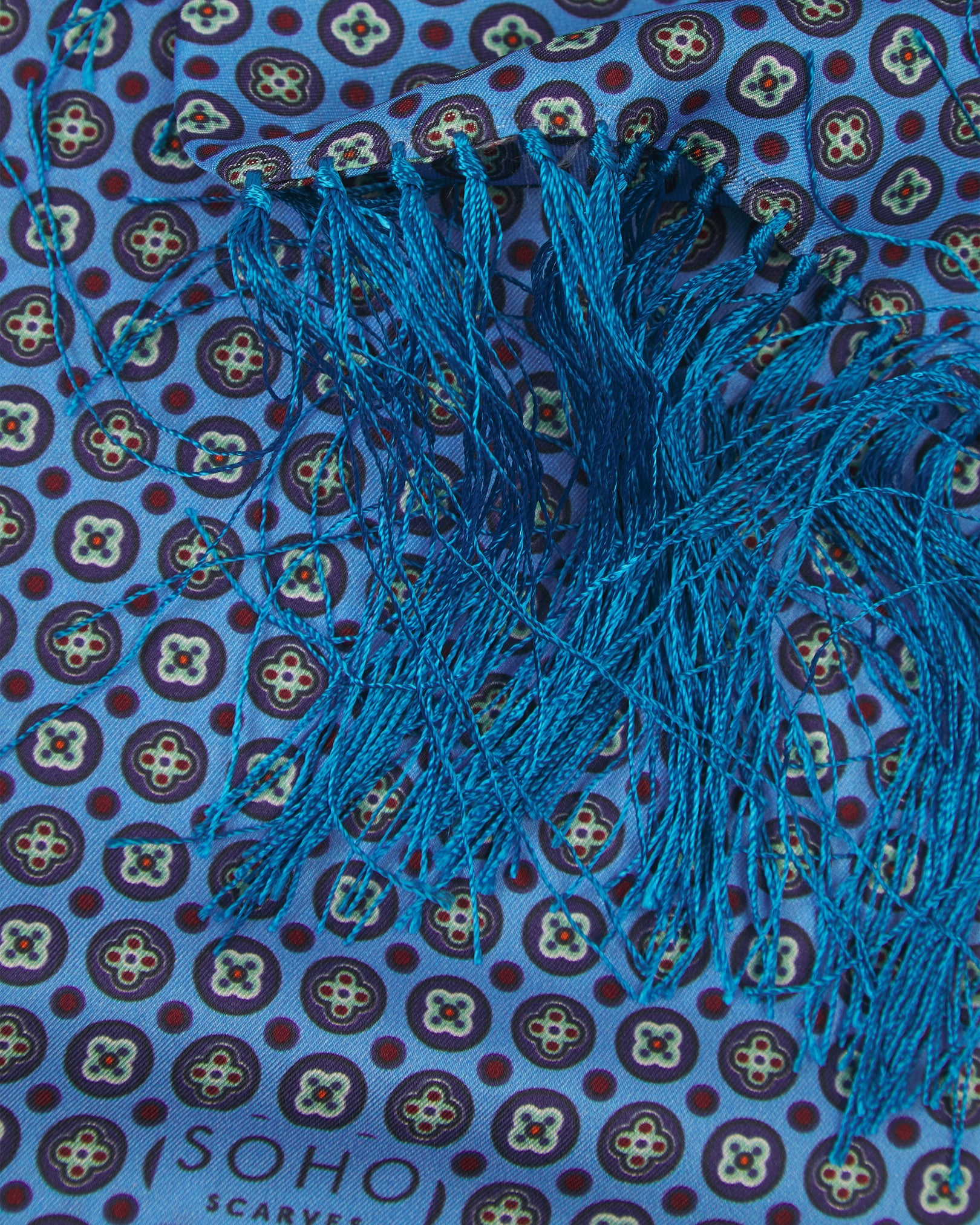 A ruffled close-up of the 'Toshima' Gardenia silk aviator scarf, presenting the overlapping elements of multicoloured stylised floral patterns patterns against a blue background.