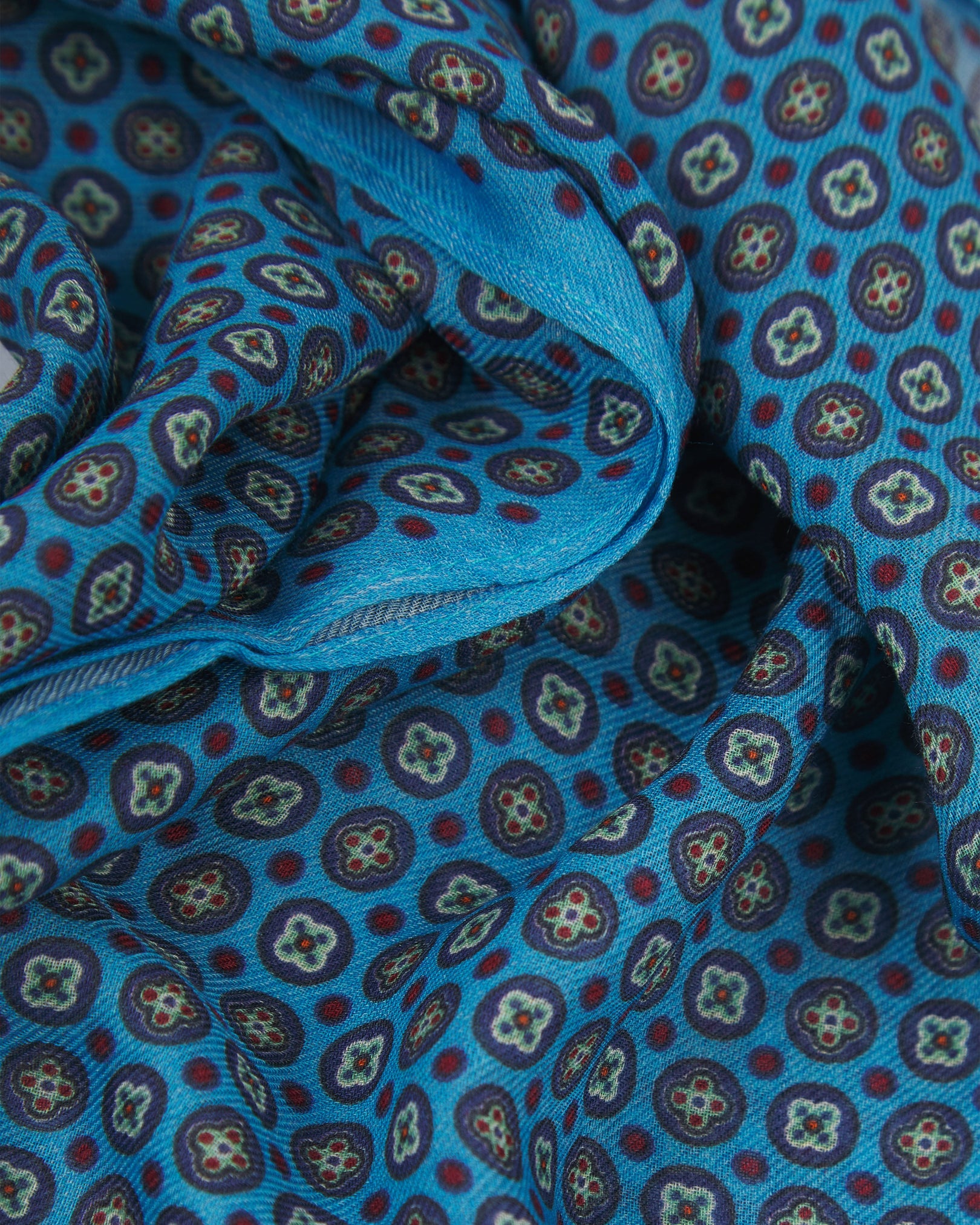 A ruffled close-up of the Toshima 'Gardenia' bohemian wide scarf, showing the soft modal and wool blend fabric with small stylised floral motifs in blue, purple, mint green and red on a light blue background.