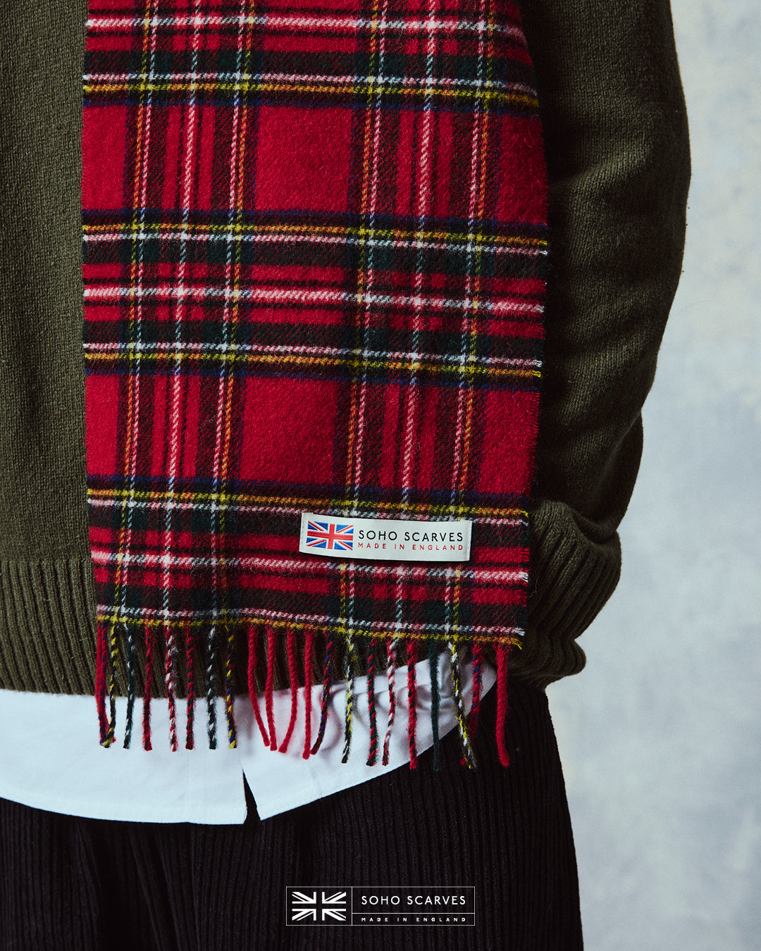 Close up of man wearing the Yorkshire-made, 'Highlands Tartan in Red' wool scarf, with focus on the 'Soho scarves, Made in England' label and matching fringes.