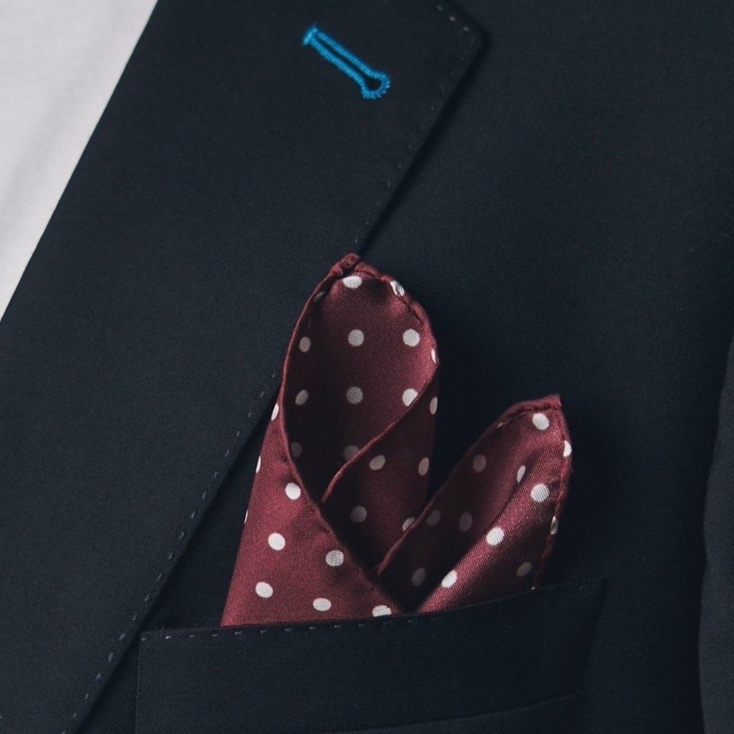 Silk square handkerchief presented in a ‘cone’ and placed in a suit jacket pocket, showing the burgundy fabric complemented by small white dots.