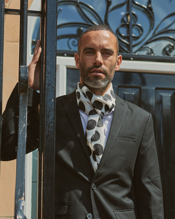 Male model wearing 'The Gable' silk aviator scarf from 'The Basics' collection. Tied loosely around neck in a simple knot and shoulders and paired with a smart, dark suit.