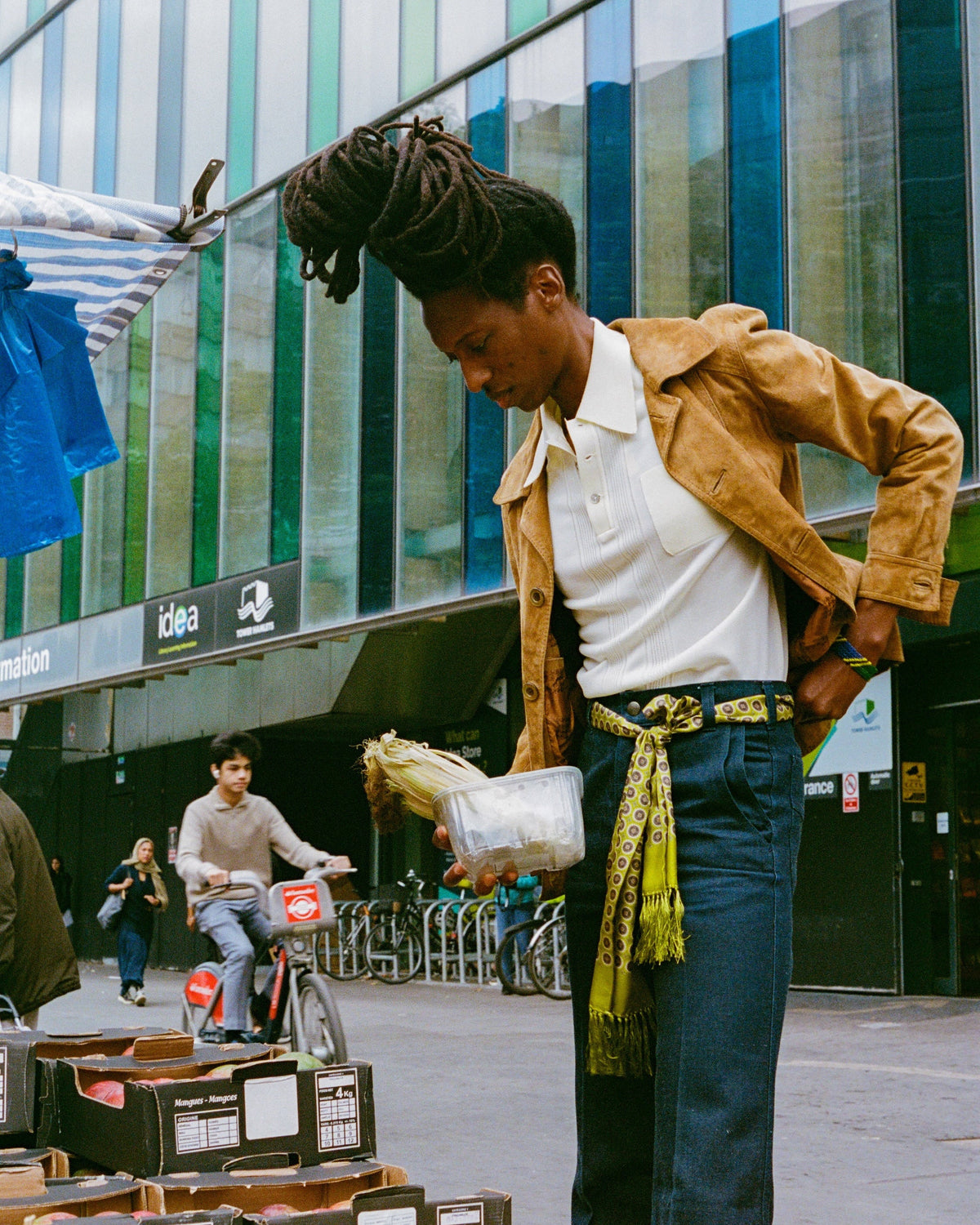 A person standing on a city street in front of a modern glass building, wearing a tan jacket over a white polo shirt, dark blue jeans, and a patterned yellow-green belt. Boxes are stacked in the foreground, and a cyclist is visible in the background.