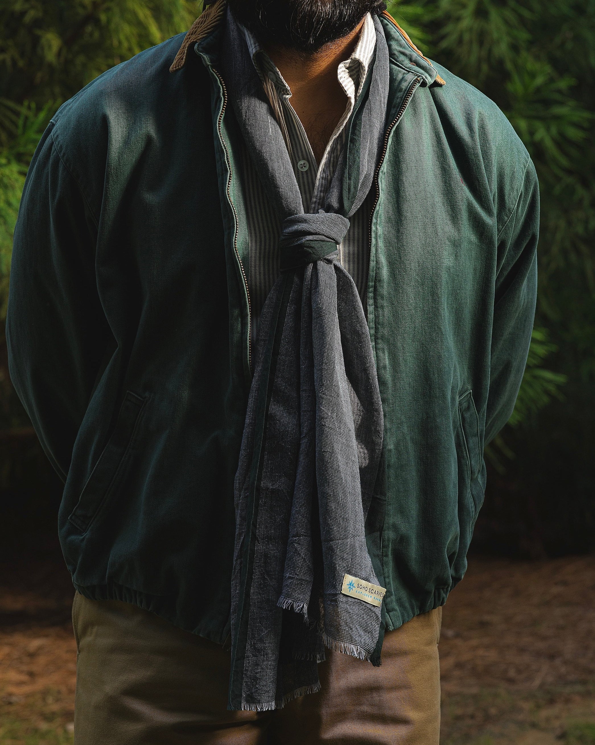 A man with a beard and glasses wearing a dark green bomber jacket, a gray knit scarf tied around his neck, and tan pants, standing outdoors in front of a lush green background.