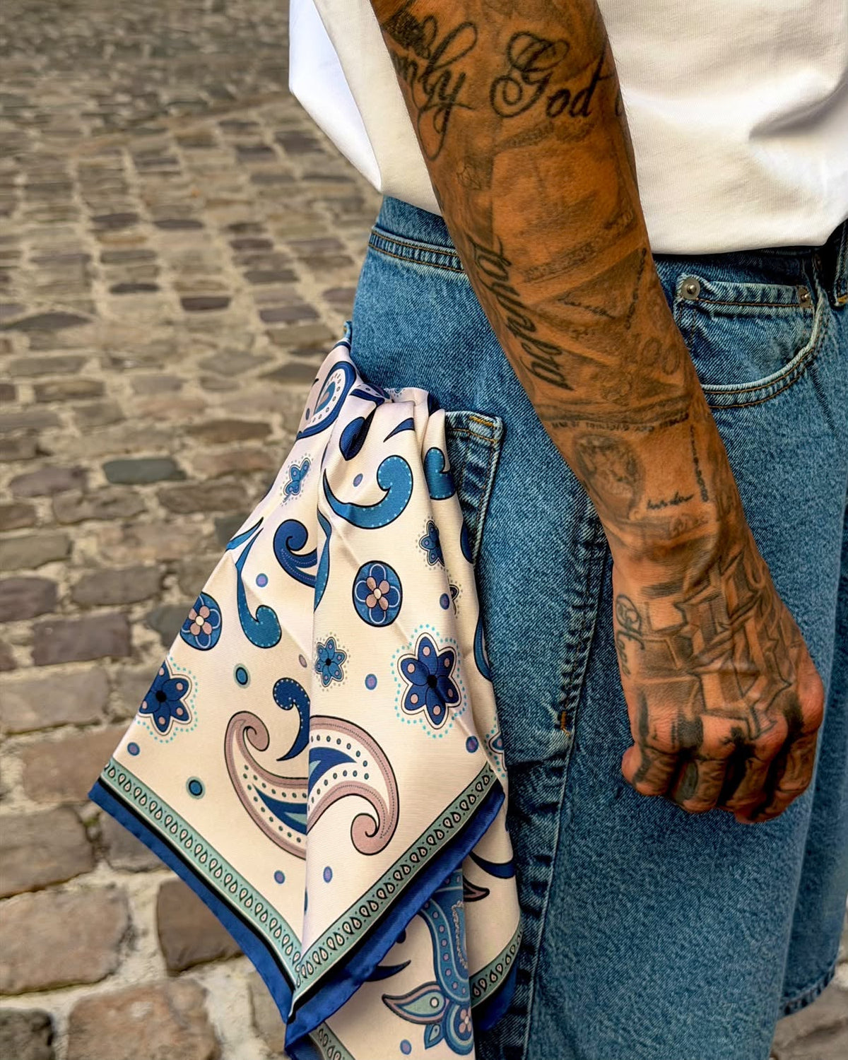 A close-up lifestyle shot of a person wearing blue denim shorts and a white t-shirt, with a cream-colored bandana featuring a blue paisley pattern tucked into the back pocket. The background is a cobblestone street.