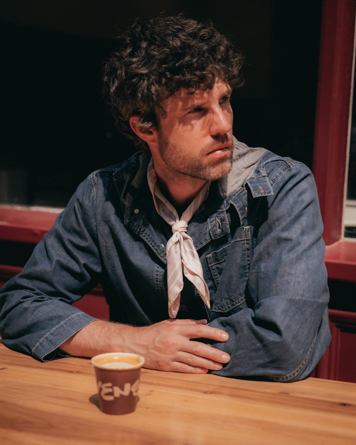 A man with curly dark hair sits at a wooden table wearing a blue denim shirt over a black top, with a light-colored necktie and a small paper cup in front of him.