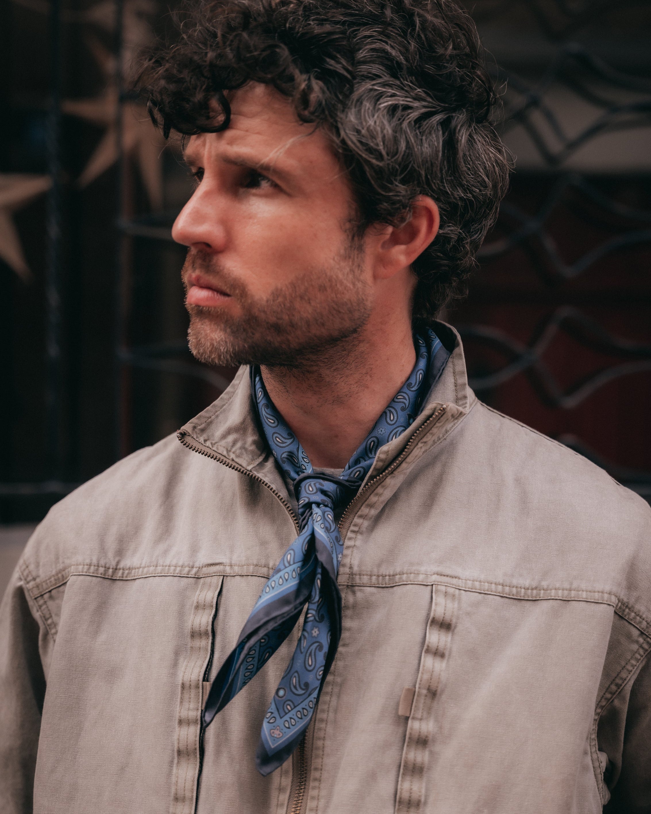 A man in profile wearing a light beige utility jacket over a blue paisley neck scarf, set against a dark, patterned background.