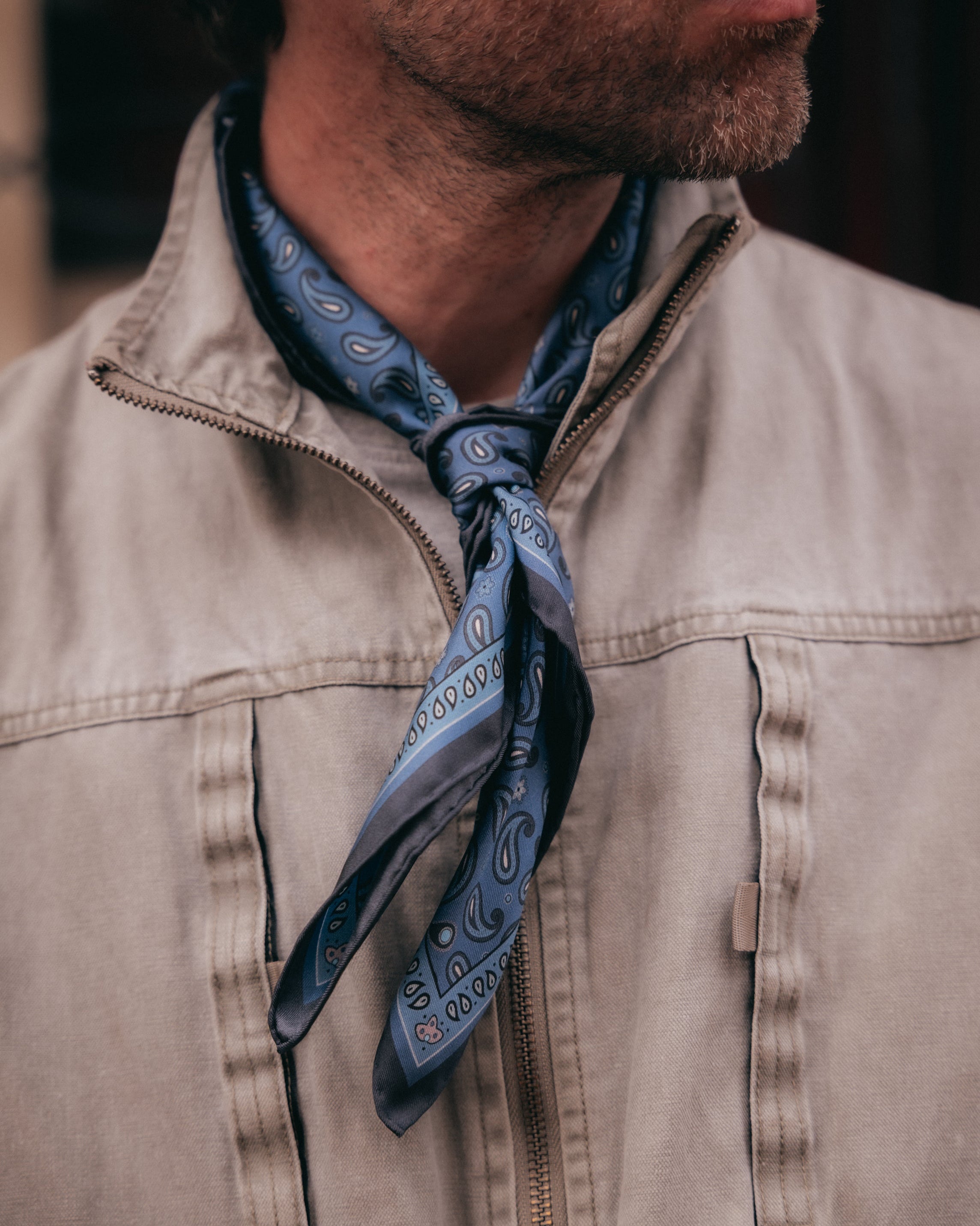 A close-up of a man wearing a beige zip-up jacket and a blue paisley neck scarf.