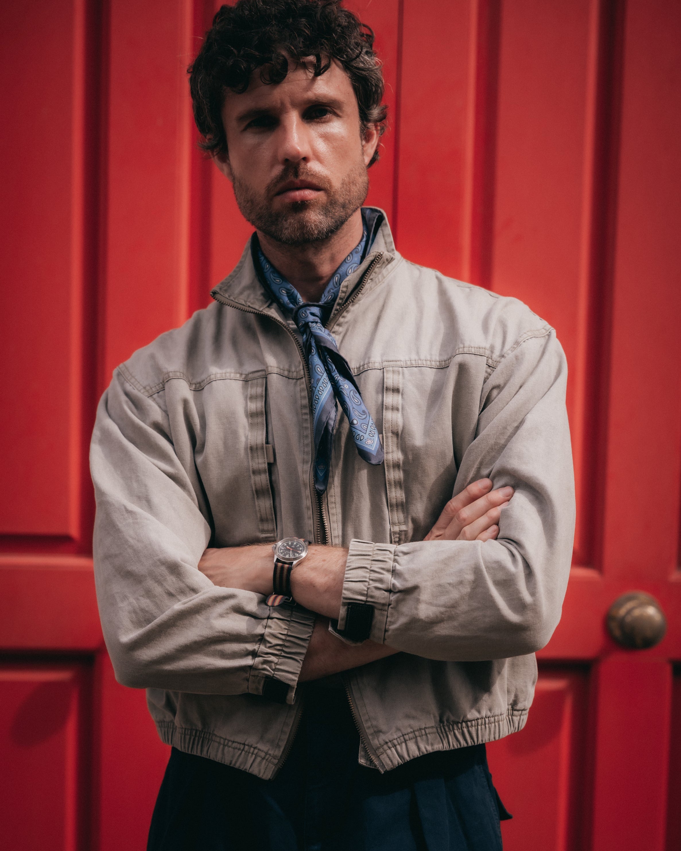A man with curly dark hair and a beard stands with arms crossed in front of a bright red paneled door. He is wearing a light beige jacket over a blue shirt with a blue neck scarf.