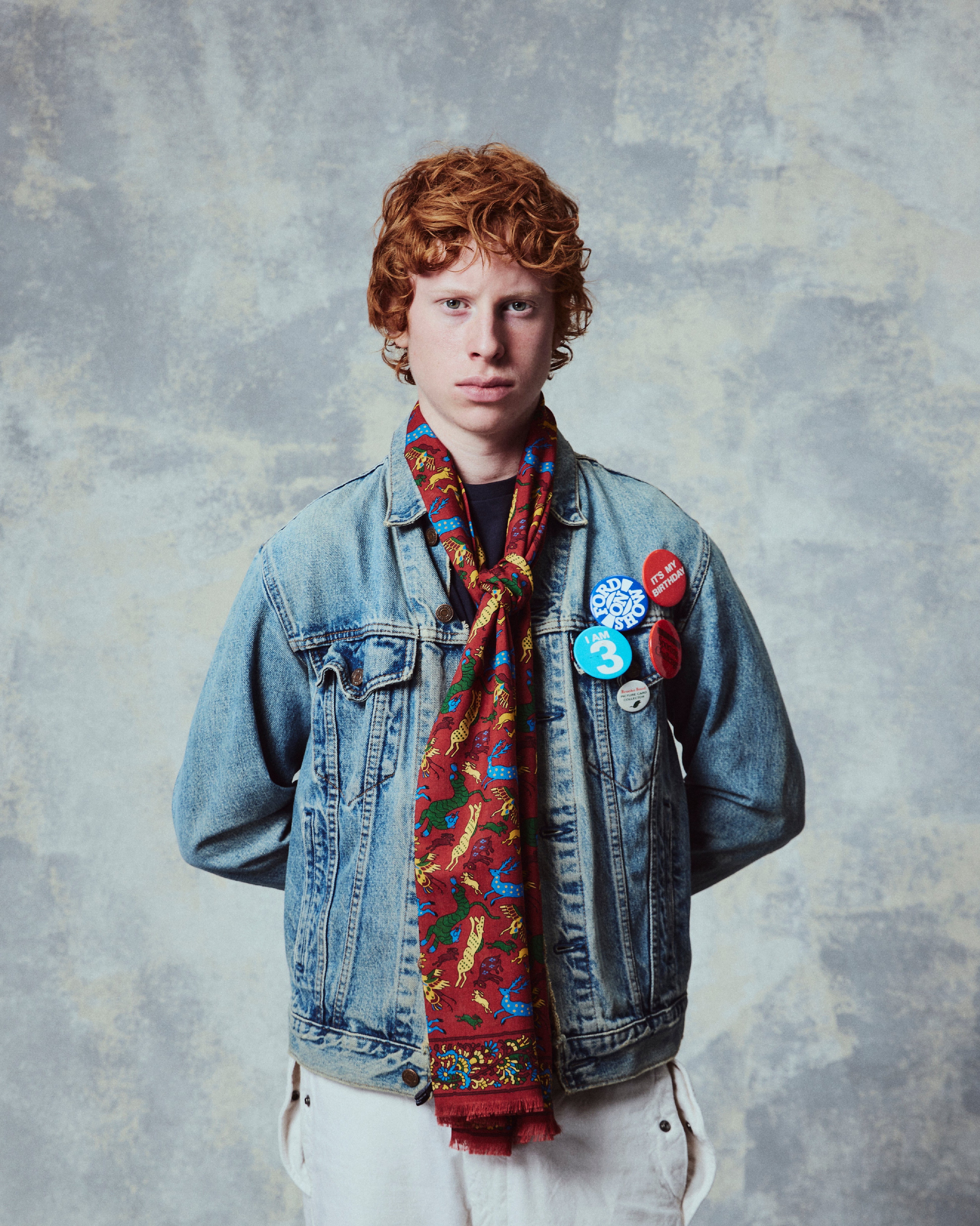 Person wearing a denim jacket, colorful scarf, and various pins on a textured gray background