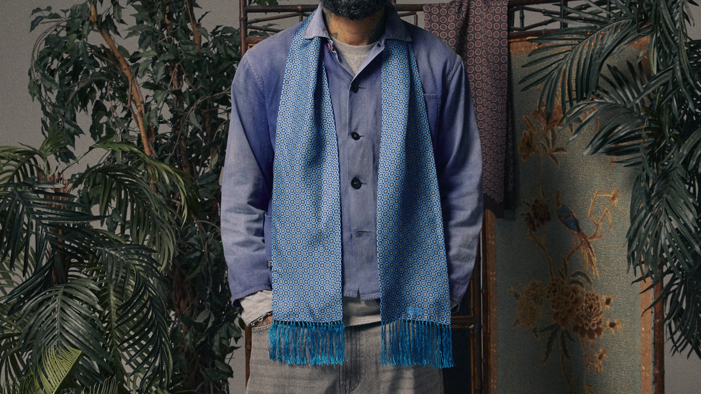 A man wearing a blue patterned scarf with fringe, a blue shirt, and gray trousers, standing in a styled room with potted plants, a folding screen, and a framed painting.