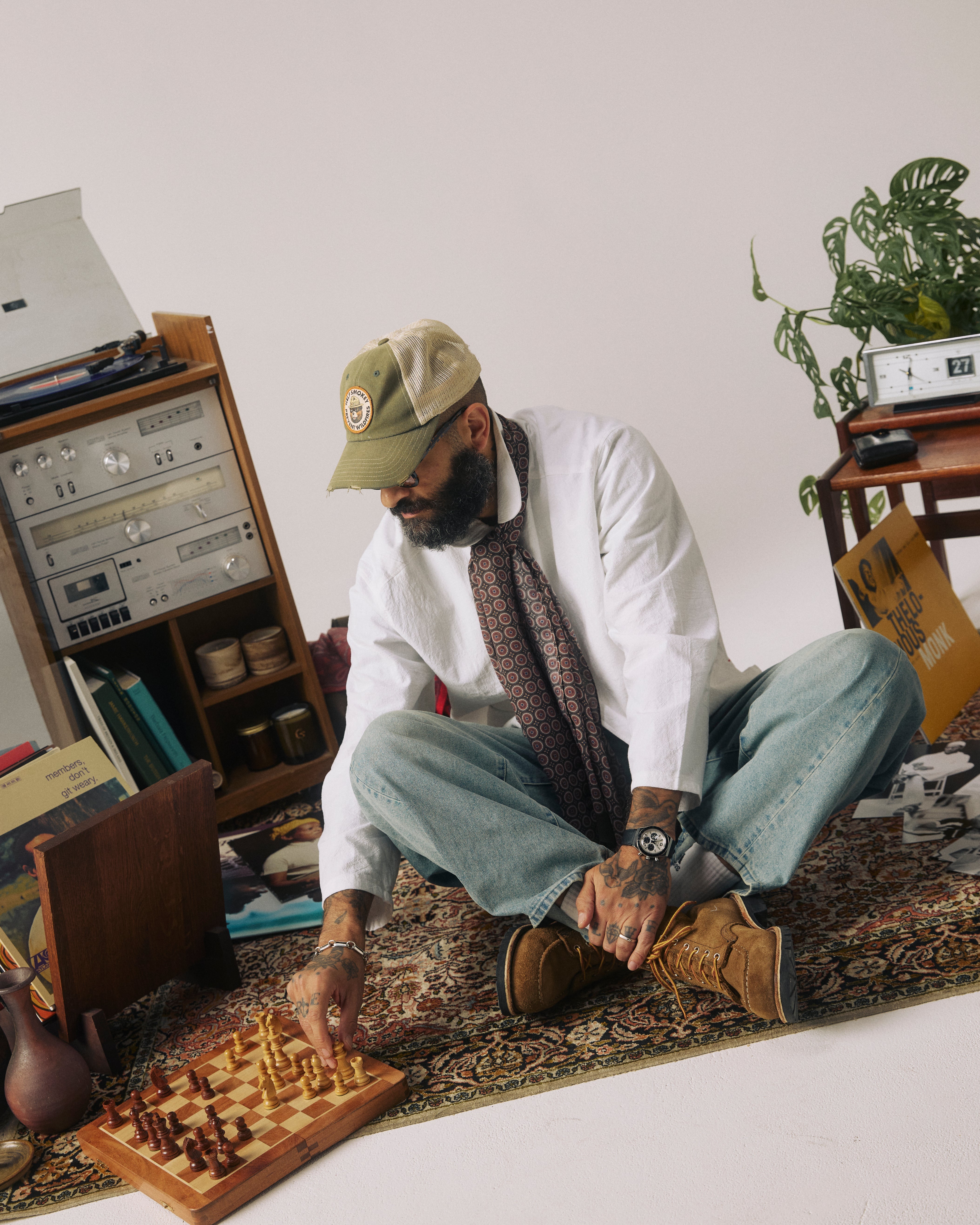 A man sits cross-legged on a patterned rug in a styled indoor room, wearing a white shirt, patterned tie, blue jeans, brown boots, and a tan baseball cap while interacting with a chessboard. A record player, books, and a potted plant are visible in the background.