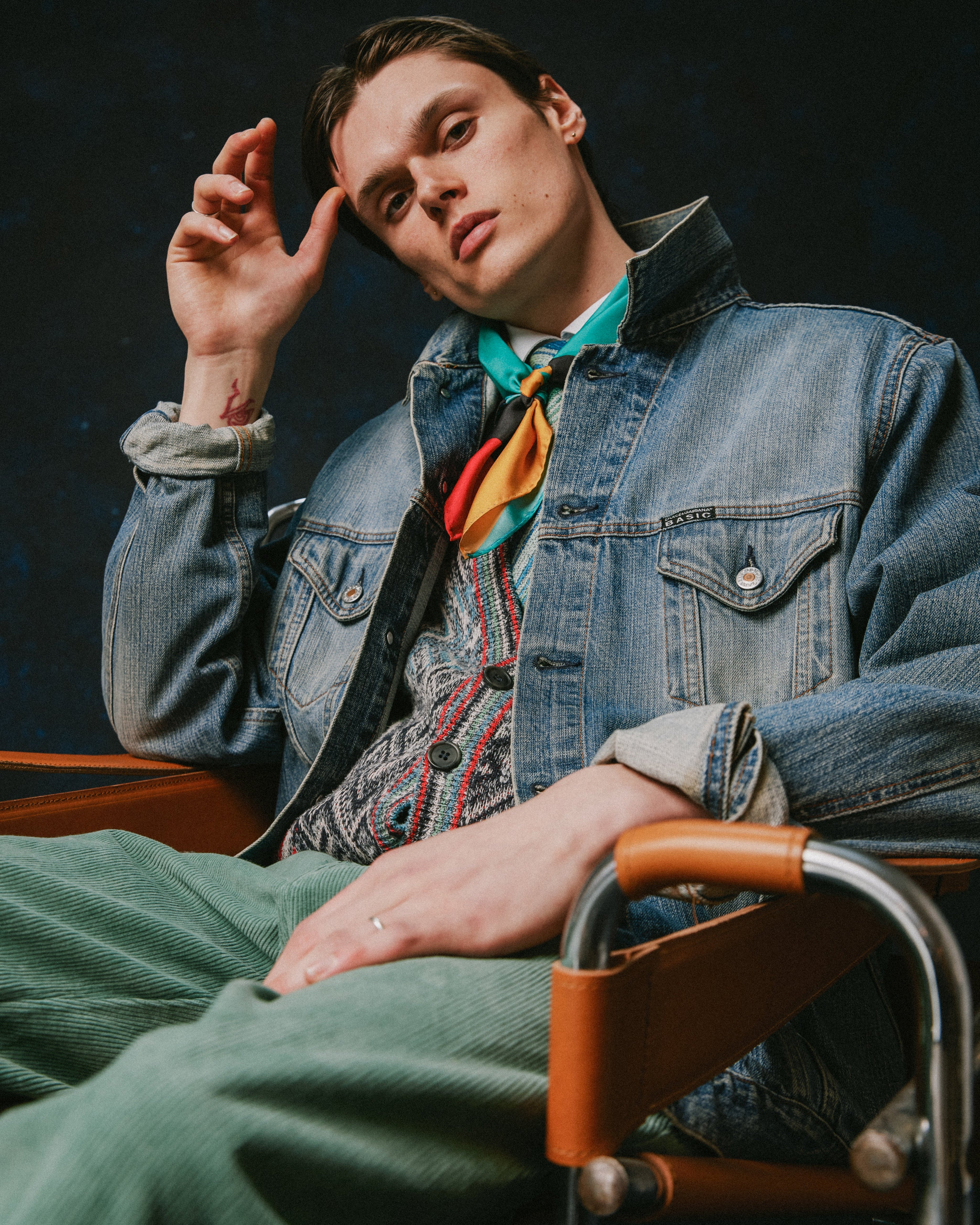 A man wearing a blue denim jacket, patterned shirt, bright turquoise and orange neck scarf, and green corduroy trousers, seated in a wooden chair against a dark studio background.