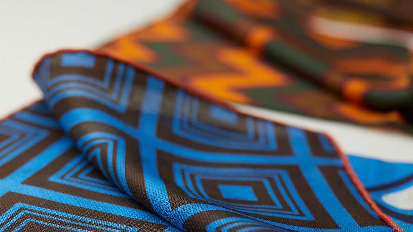 A close-up of a folded patterned fabric or scarf with a bold geometric design in bright blue, dark brown, and orange, set against a clean white background.