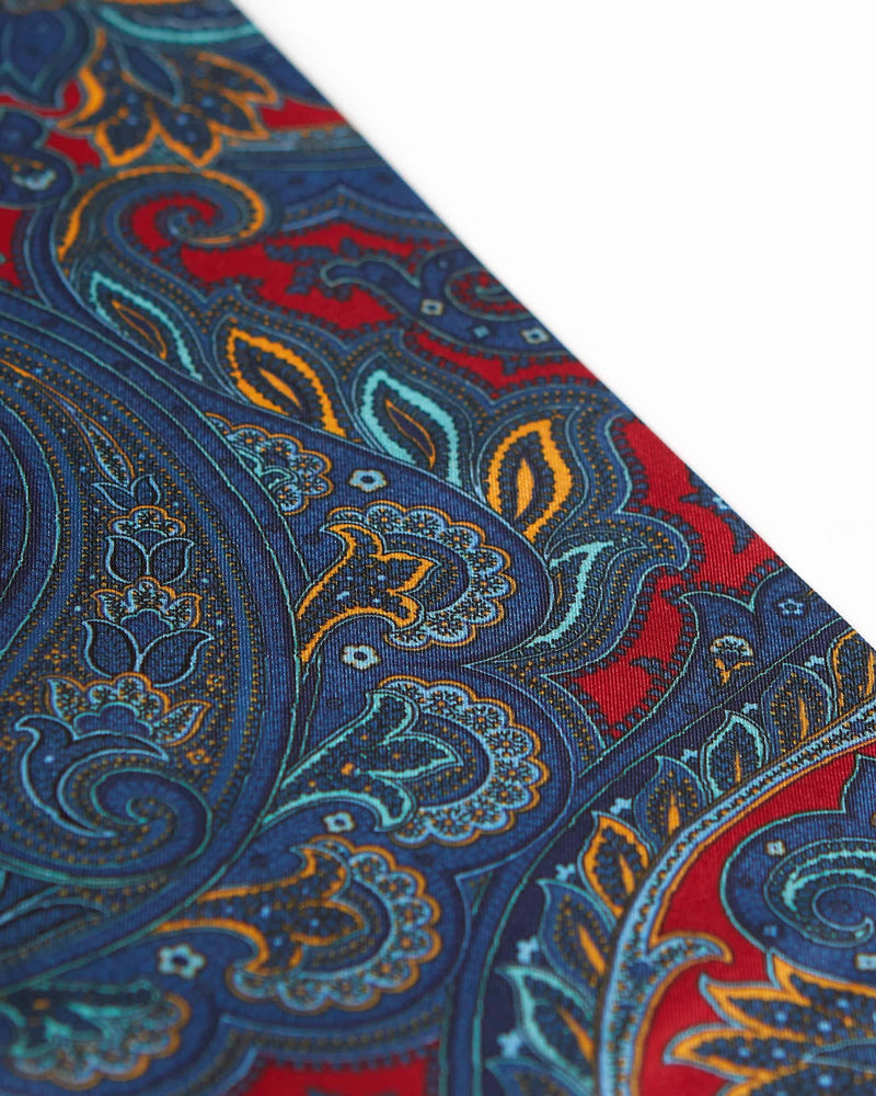 Angled view of the light and dark blue, orange and grey paisley patterns on the deep red scarf.
