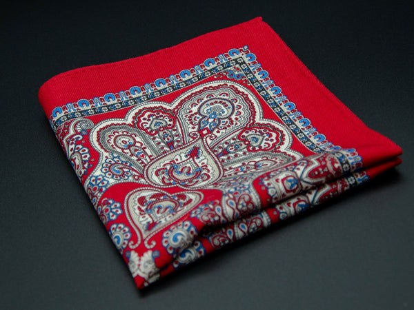 Pure wool 'Pegulin' pocket square folded into a quarter, clearly showing the heart-shaped paisley motifs and ornate blue border on a bright red ground.