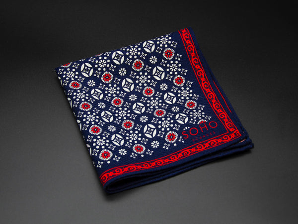 Wool 'Ooty' pocket square folded showing white and red decorative floral patterns aranged in linear repeats on a navy ground. Framed by an intricate red border. SOHO Scarves branding clearly visible on bottom-right.